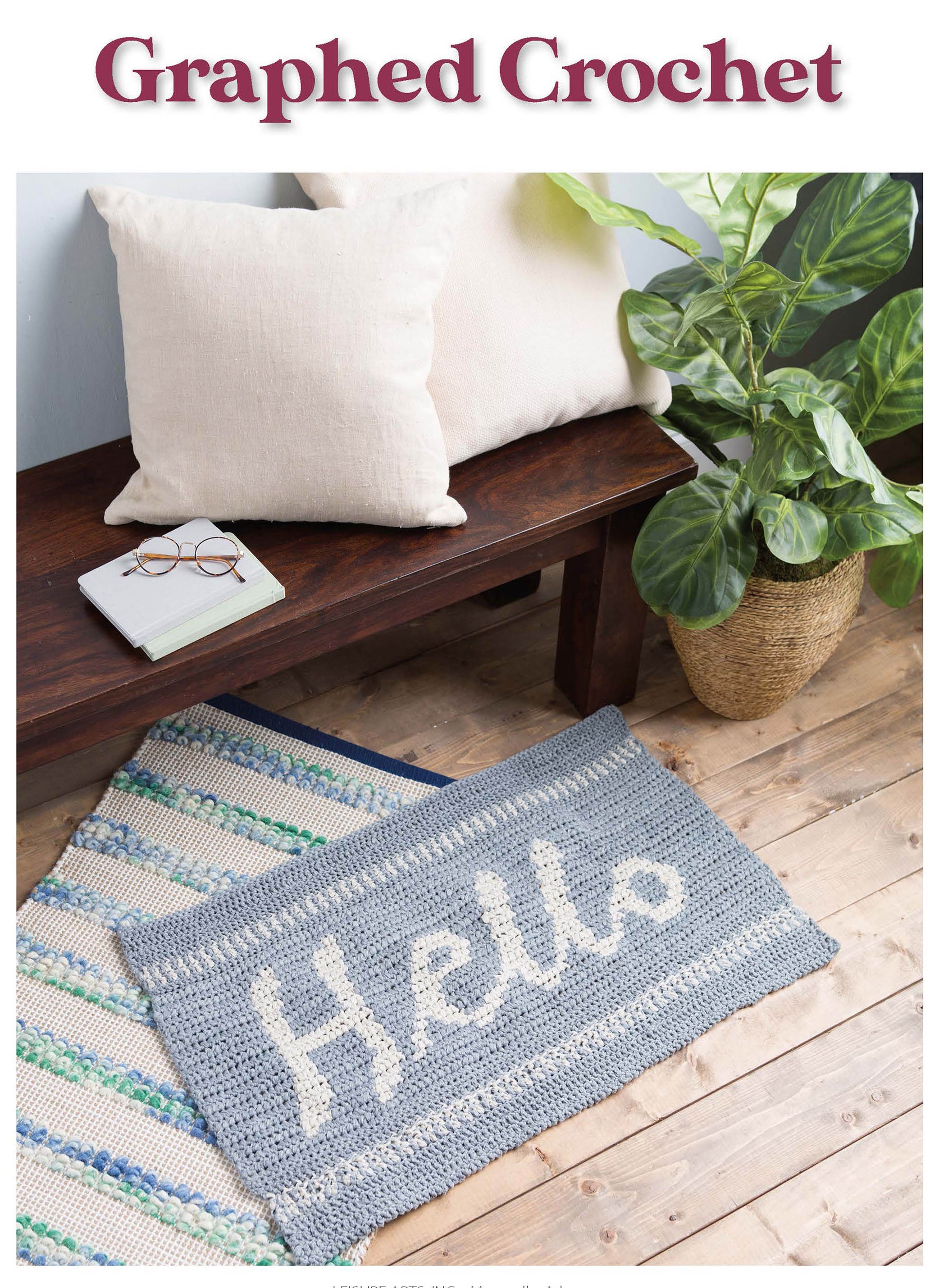 The Graphed Crochet Digital Download by Leisure Arts features a gray mat with “Hello” in white yarn, perfect for cozy home décor. Shown on a wooden floor by a blue-striped rug, bench, cream cushions, and leafy plant.
