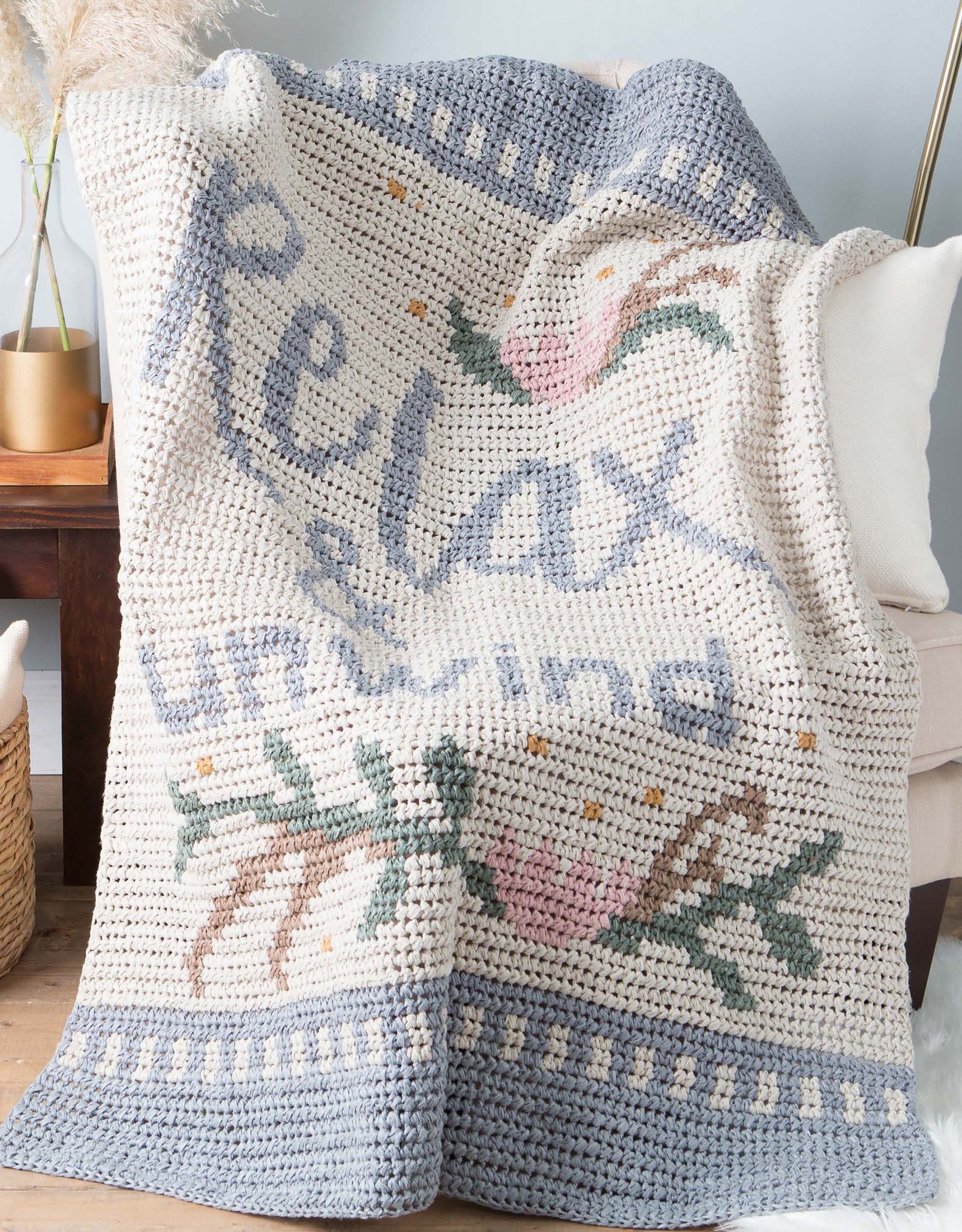 The Graphed Crochet Digital Download by Leisure Arts features a blue “Relax, Unwind” message with pink flowers, green leaves, and a gray border—a cozy home accent—shown draped over a white cushion on a chair.