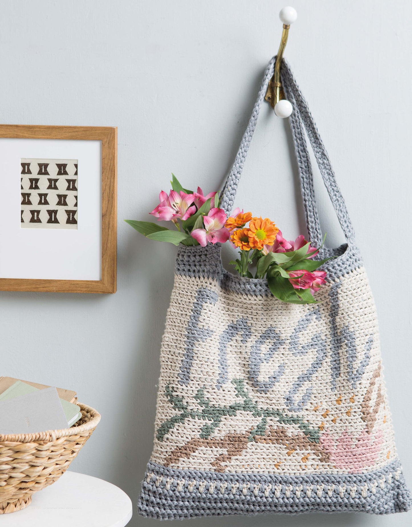 A Graphed Crochet Digital Download by Leisure Arts hangs on a wall hook, adorned with the word "fresh" and florals, brimming with colorful flowers. Nearby are a framed print and a wicker basket of envelopes on a white surface.