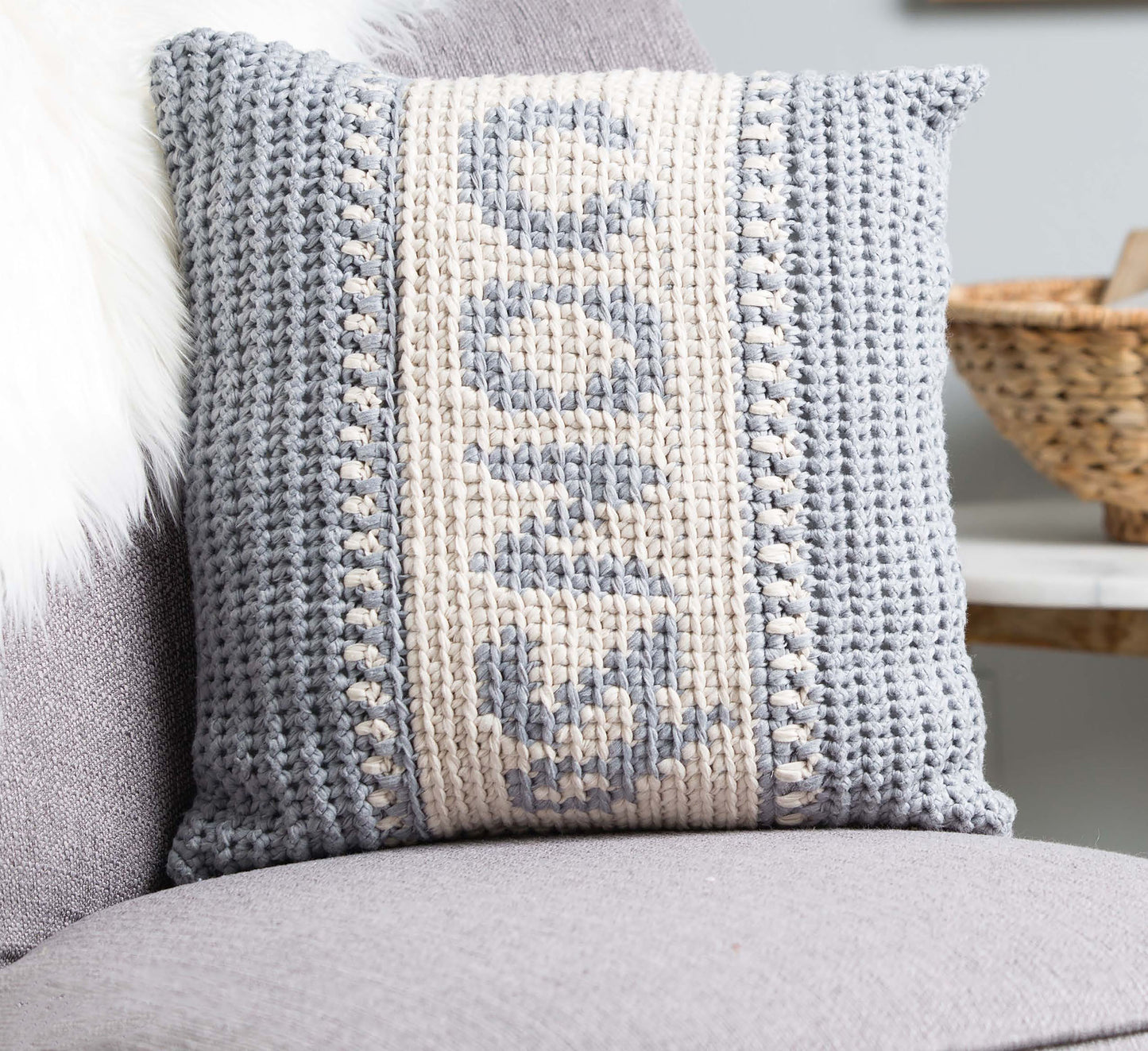 A light gray and cream crocheted pillow made with the Graphed Crochet Digital Download by Leisure Arts sits on a gray chair, featuring “SNUG” and cozy texture—perfect for warm home décor. A woven basket and white fur appear in the background.