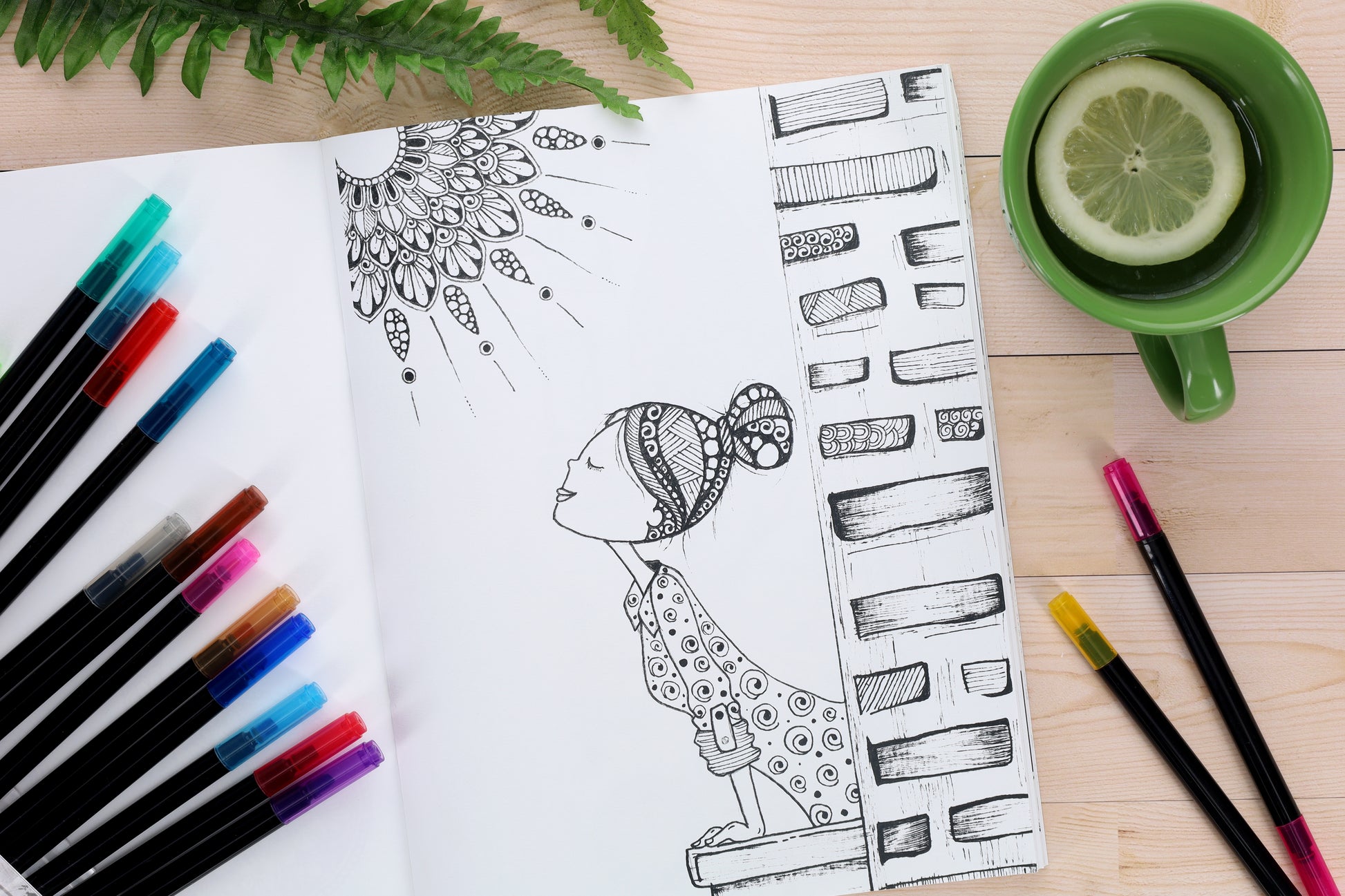 A Leisure Arts "Small Joys of Life" sketchbook featuring a black ink drawing of a woman looking up at a stylized sun, surrounded by colored pens, green lemon tea, and a fern on a wooden table—simple creative bliss.