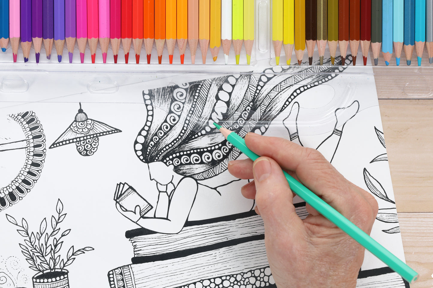 A hand colors a detailed black-and-white drawing from Small Joys of Life by Leisure Arts, adding teal to a woman’s patterned hair. Colored pencils are arranged above, highlighting the creative joy of coloring.