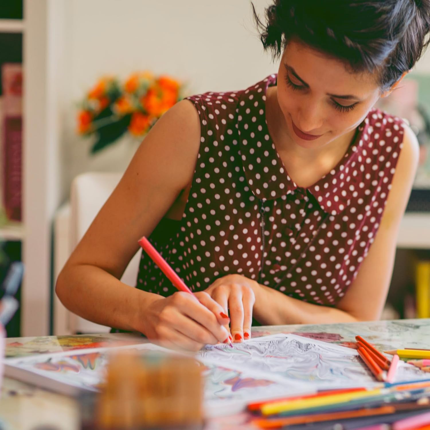 A woman in a sleeveless maroon polka-dot dress colors pages from Leisure Arts’ "Small Joys of Life" adult coloring book with colored pencils at a table, while a vase of orange flowers is blurred in the background.