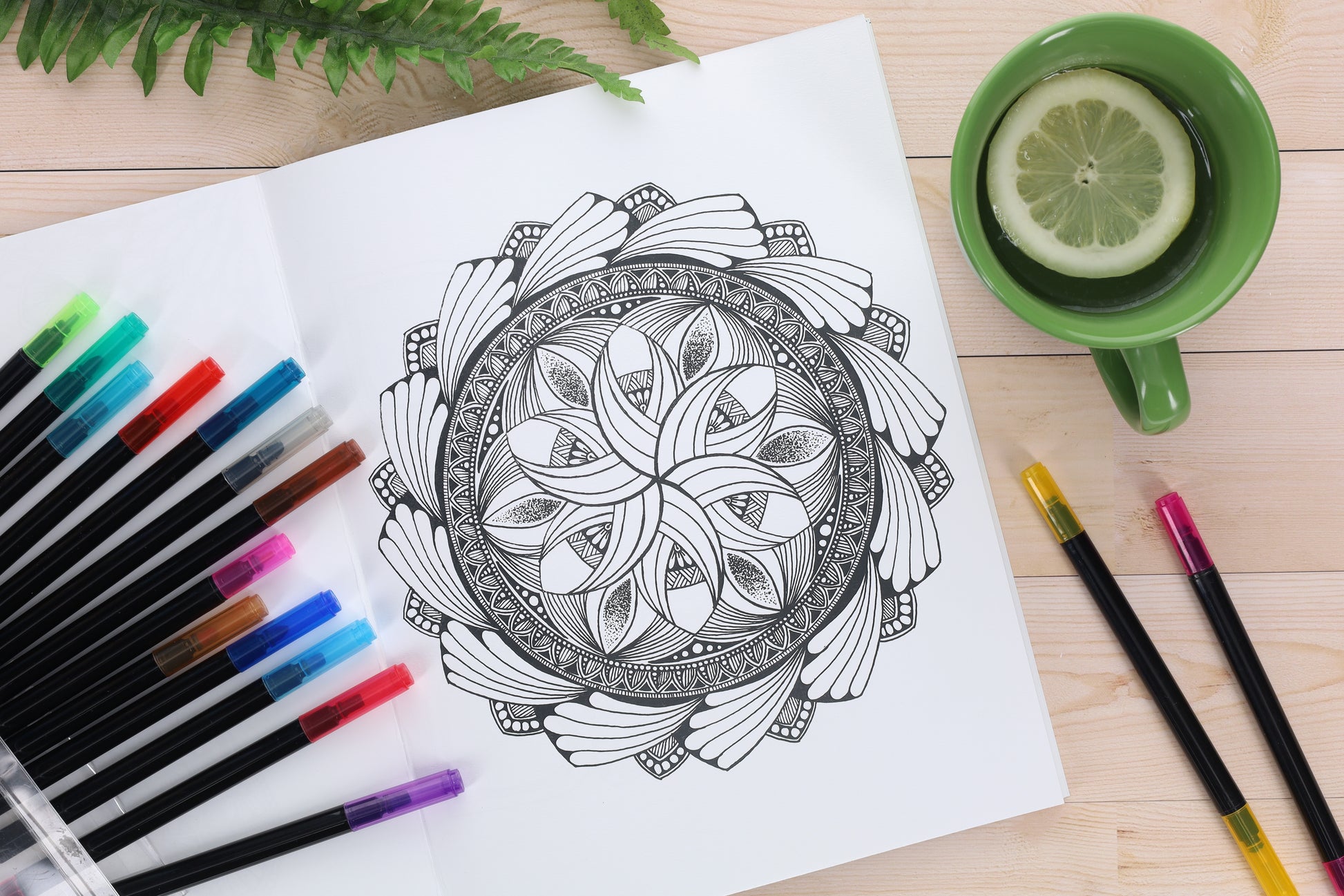 A black and white mandala page from the Meraki Mandalas Coloring Book by Leisure Arts is surrounded by colorful pens. A green cup with lemon water sits on the wood table, with a fern partly visible above the notebook.