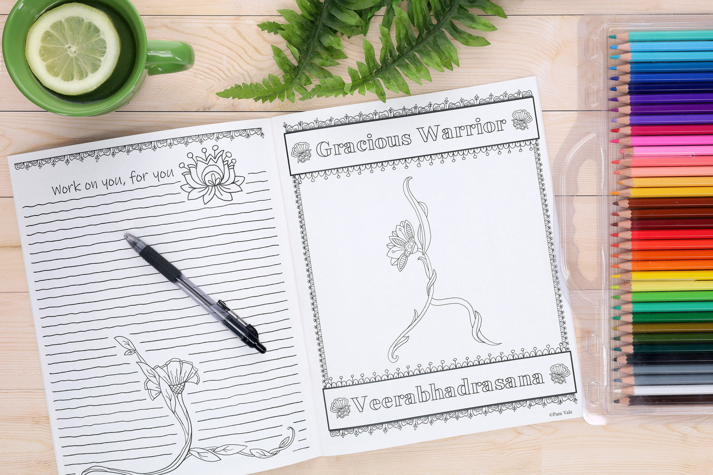 The Leisure Arts Flower Yoga Coloring Book is open to a floral Veerabhadrasana pose. Next to it are a journal, pen, colored pencils, plant, and lemon mug—capturing a calm and creative moment.