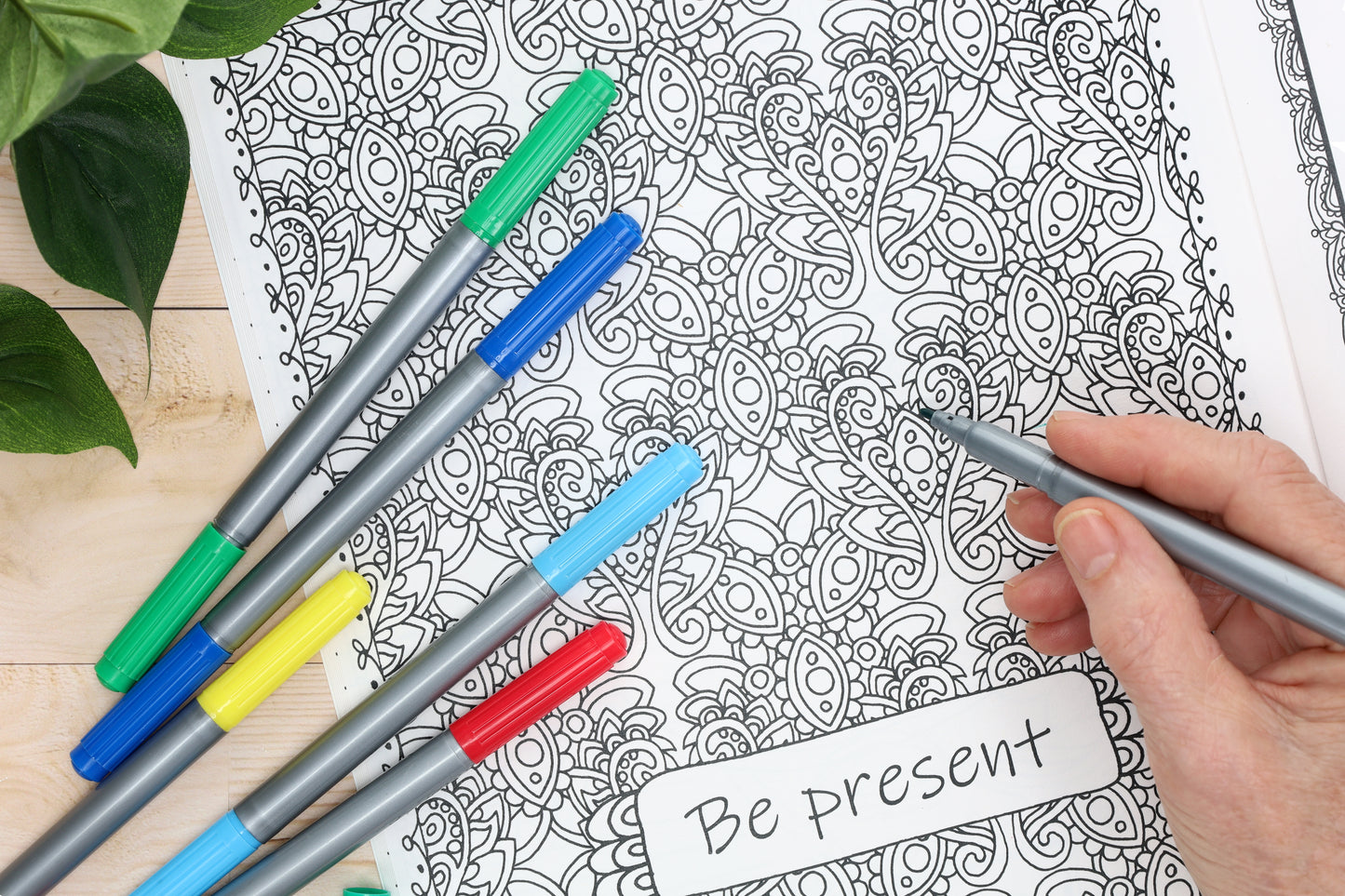 A hand holds a gray marker over the Flower Yoga Coloring Book by Leisure Arts, with colorful markers nearby and a card reading "Be present" for mindful inspiration. Green plant leaves are visible in the corner.