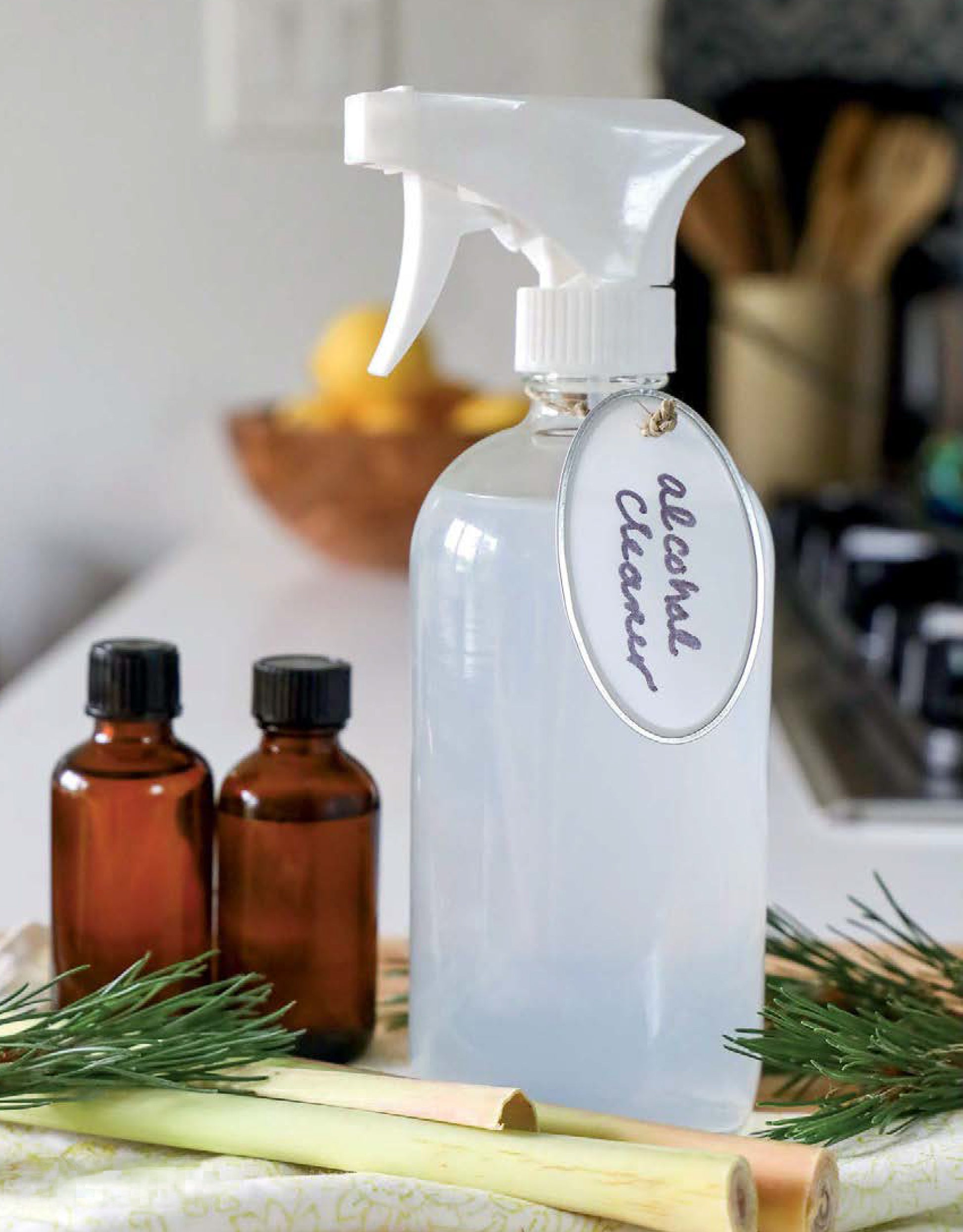 A spray bottle and brown bottles sit on a kitchen counter with pine needles, lemongrass, lemons, and utensils—perfect essentials from Leisure Arts’ Handmade Soaps, Sanitizers, & Cleaners Digital Download to help sanitize your home.