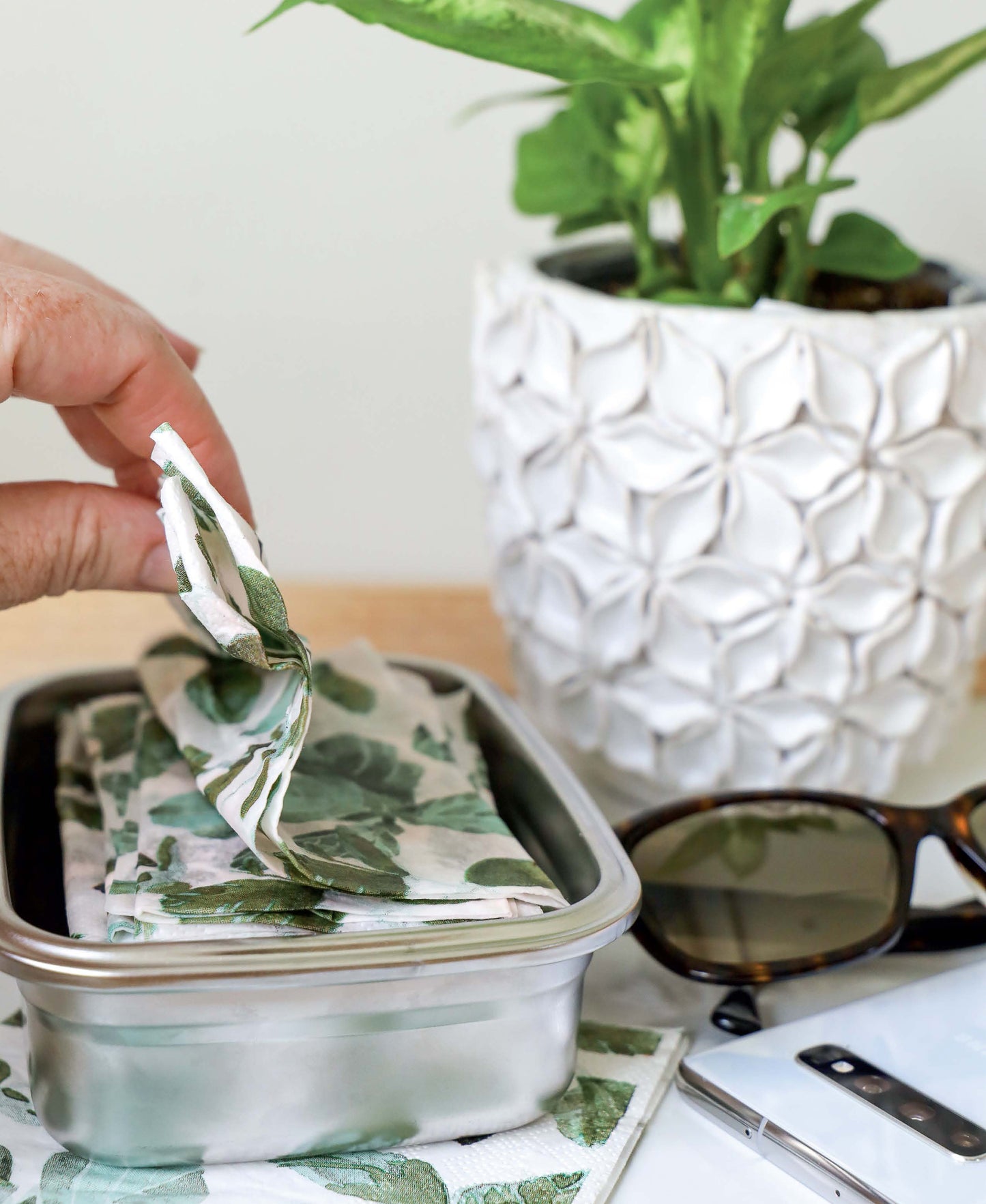 A hand displays the Leisure Arts Handmade Soaps, Sanitizers, & Cleaners Digital Download—ideal for home sanitizing. Nearby are a white plant pot with greenery, sunglasses, and a smartphone on a wooden surface.
