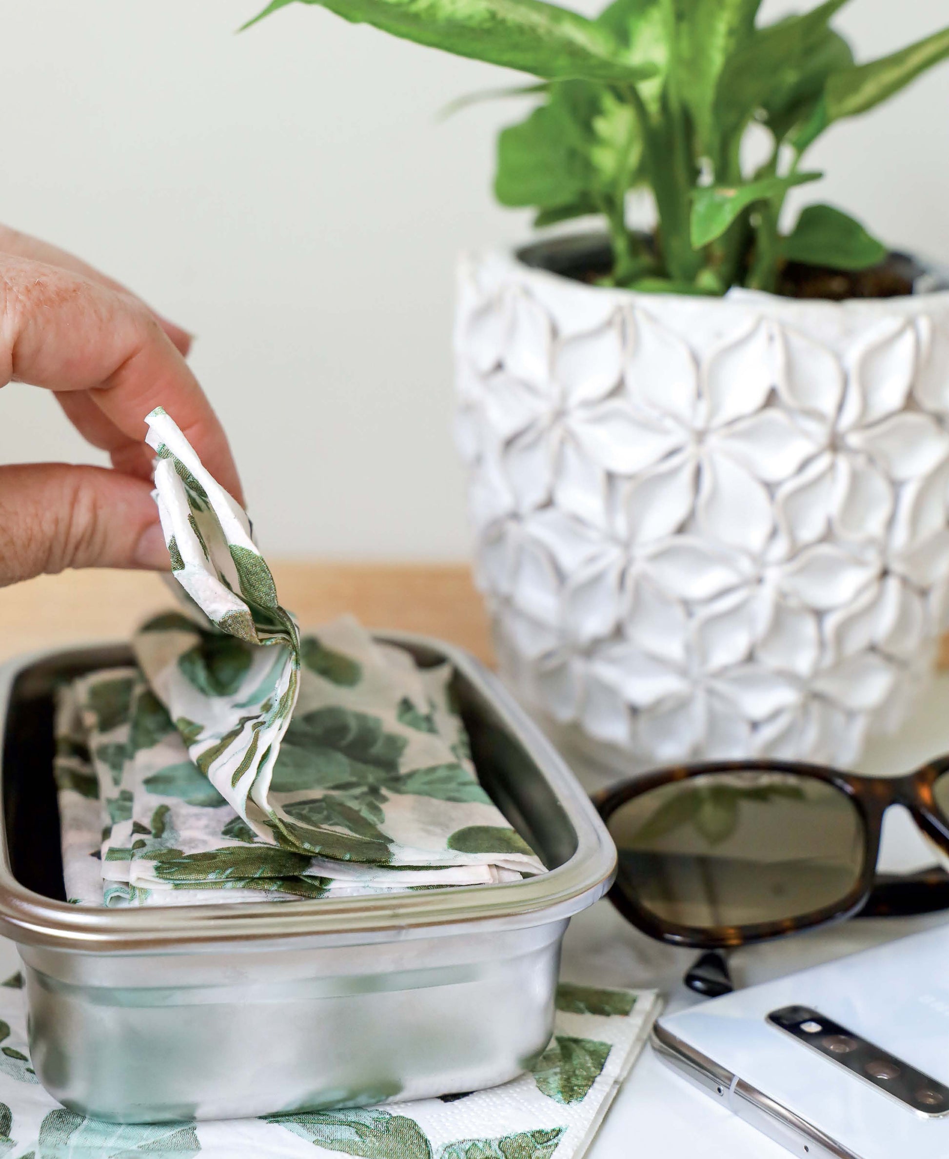 A hand displays the Leisure Arts Handmade Soaps, Sanitizers, & Cleaners Digital Download—ideal for home sanitizing. Nearby are a white plant pot with greenery, sunglasses, and a smartphone on a wooden surface.