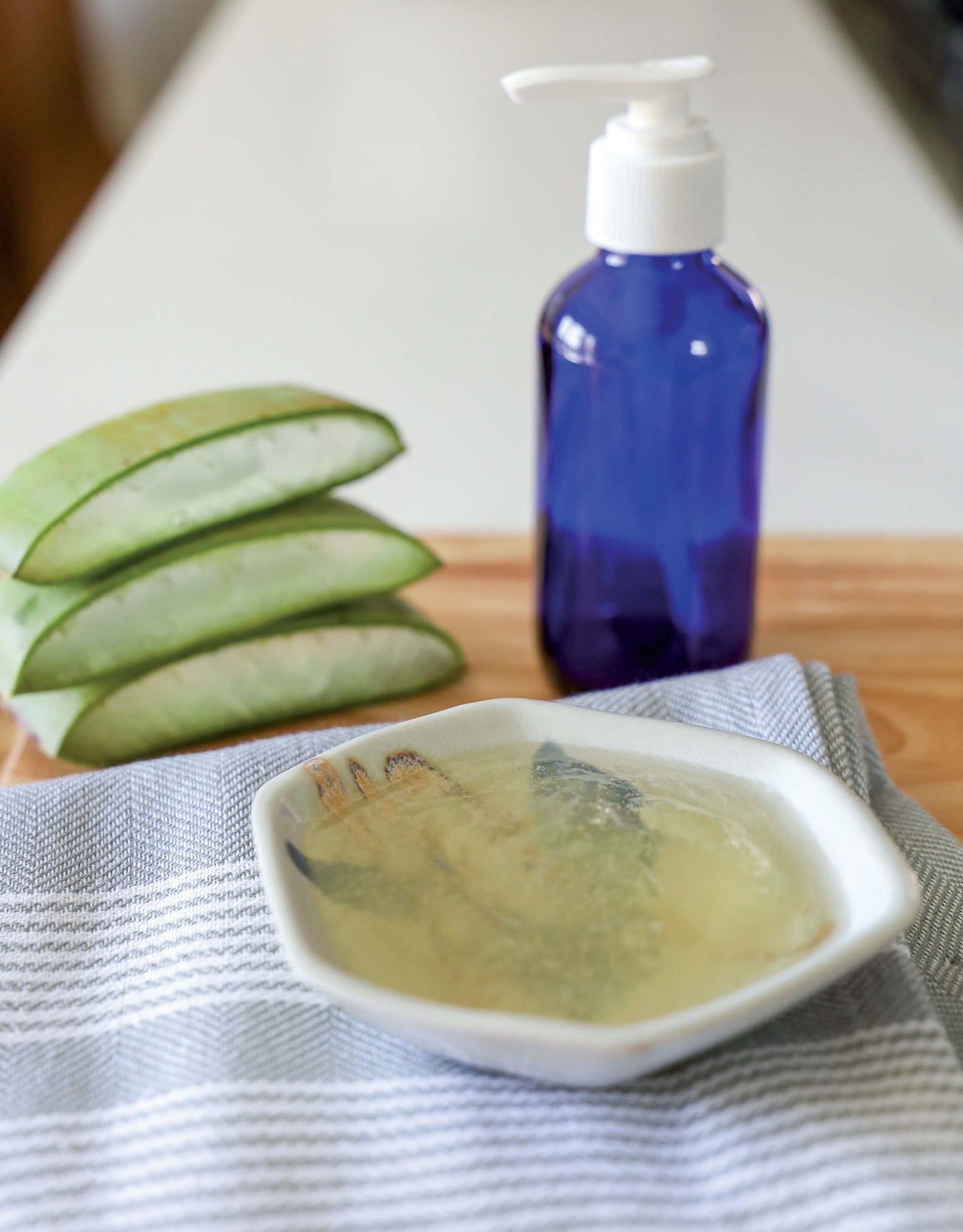 A bowl of aloe gel, sliced aloe leaves, a blue pump bottle, and hand sanitizer spray are displayed on wood—perfect for making cleaning products from the Leisure Arts Handmade Soaps, Sanitizers, & Cleaners Digital Download.