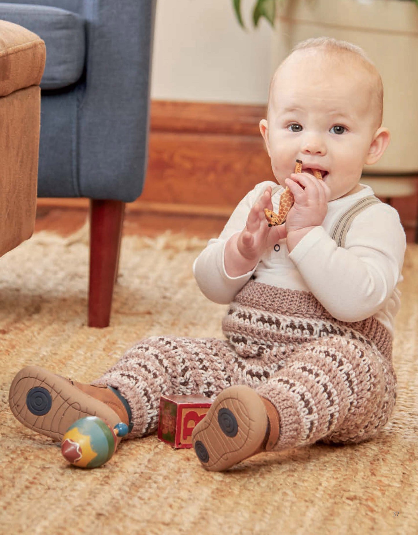 A baby in crocheted pants from Vintage Baby Crochet by Leisure Arts and a light sweater sits on a carpeted floor, smiling and holding a giraffe toy. Colorful toys and a wooden block are scattered nearby.
