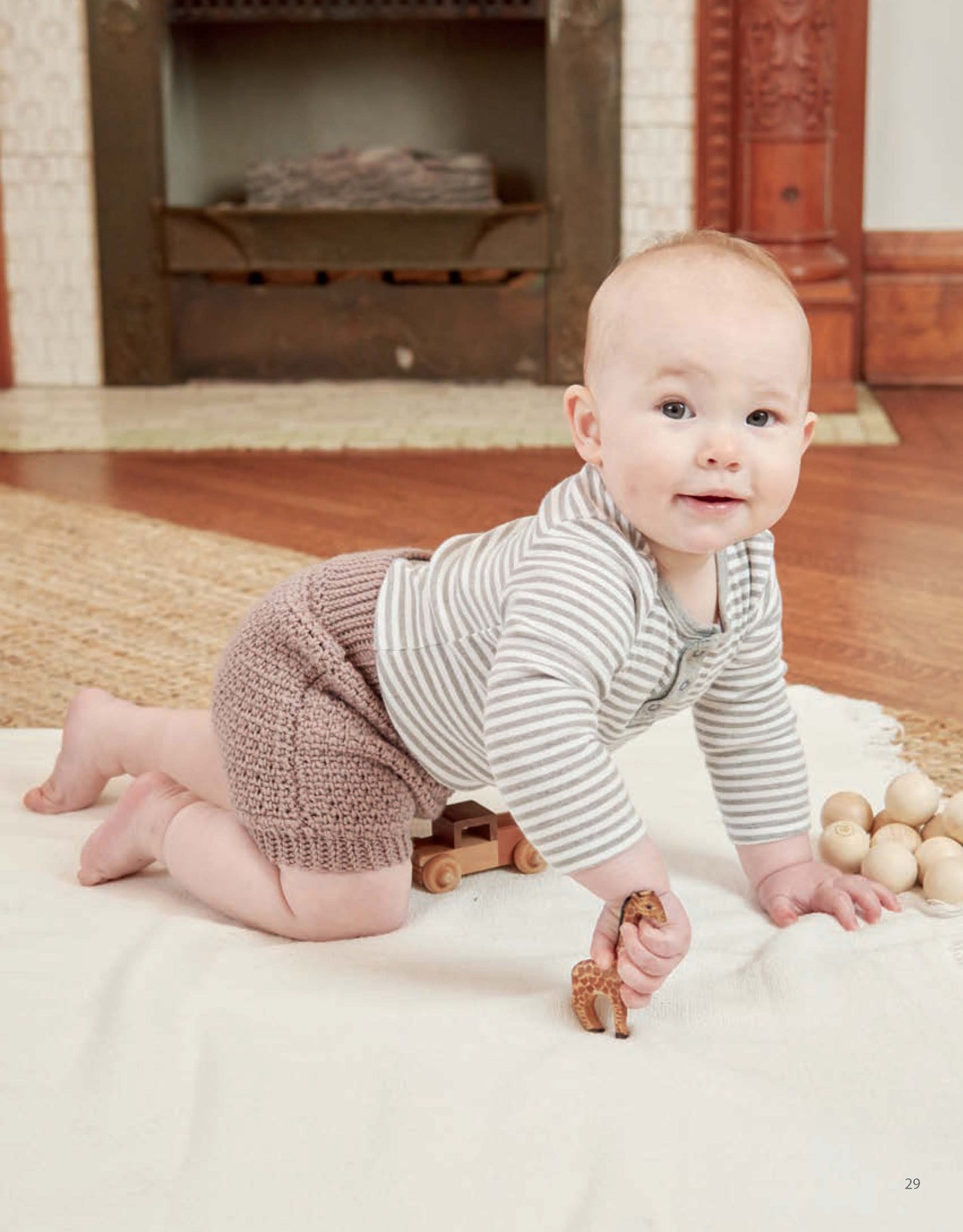 A baby in a striped shirt and knit shorts crawls on a white blanket, holding a small toy animal, with wooden toys nearby and cozy Vintage Baby Crochet patterns by Leisure Arts adding charm near the fireplace.
