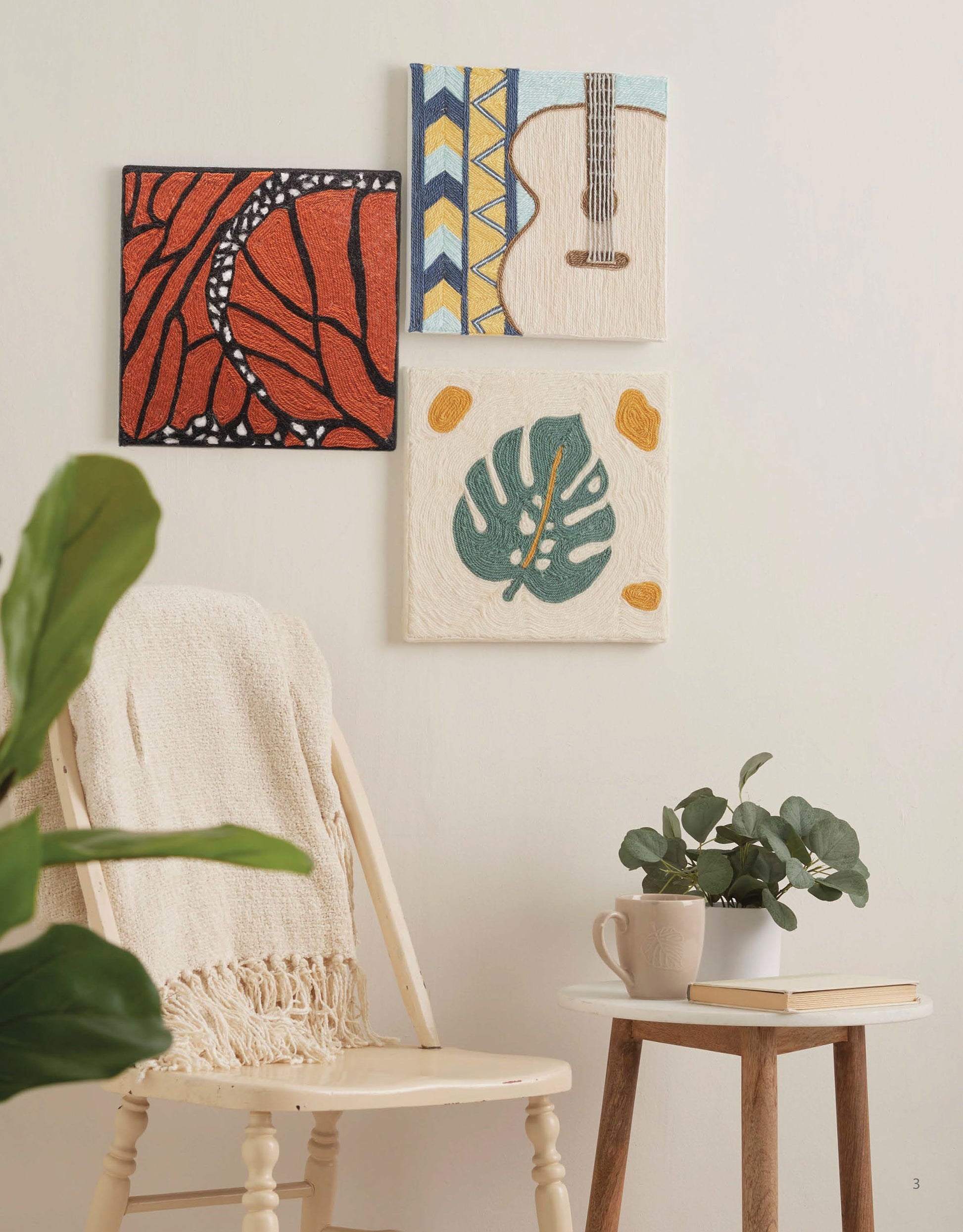 A cozy nook with a cream chair, textured blanket, small table with mug and plant, and three vibrant wall arts including the Yarn Painting Digital Download by Leisure Arts—featuring a guitar, abstract art, and a green leaf.
