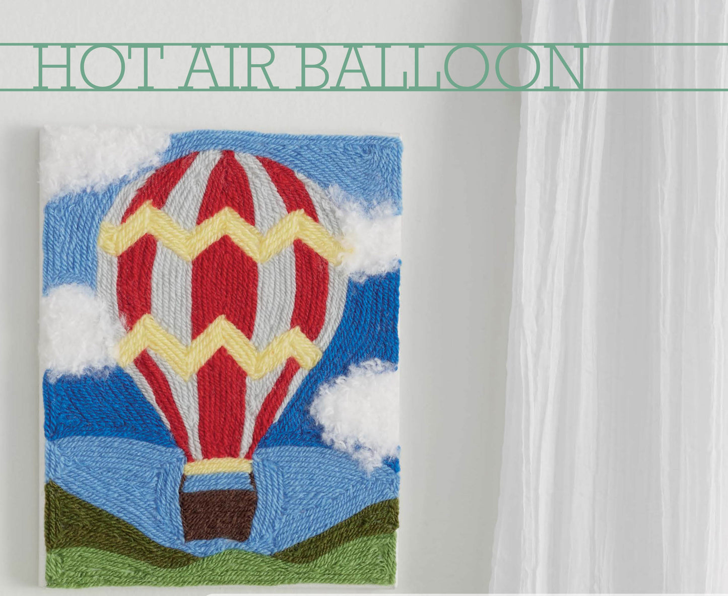 The Yarn Painting Digital Download by Leisure Arts features a hot air balloon with red, yellow, and gray stripes floating above green hills—ideal for beginners or creative canvas projects. Displayed on a white wall near sheer curtains. Text reads HOT AIR BALLOON.