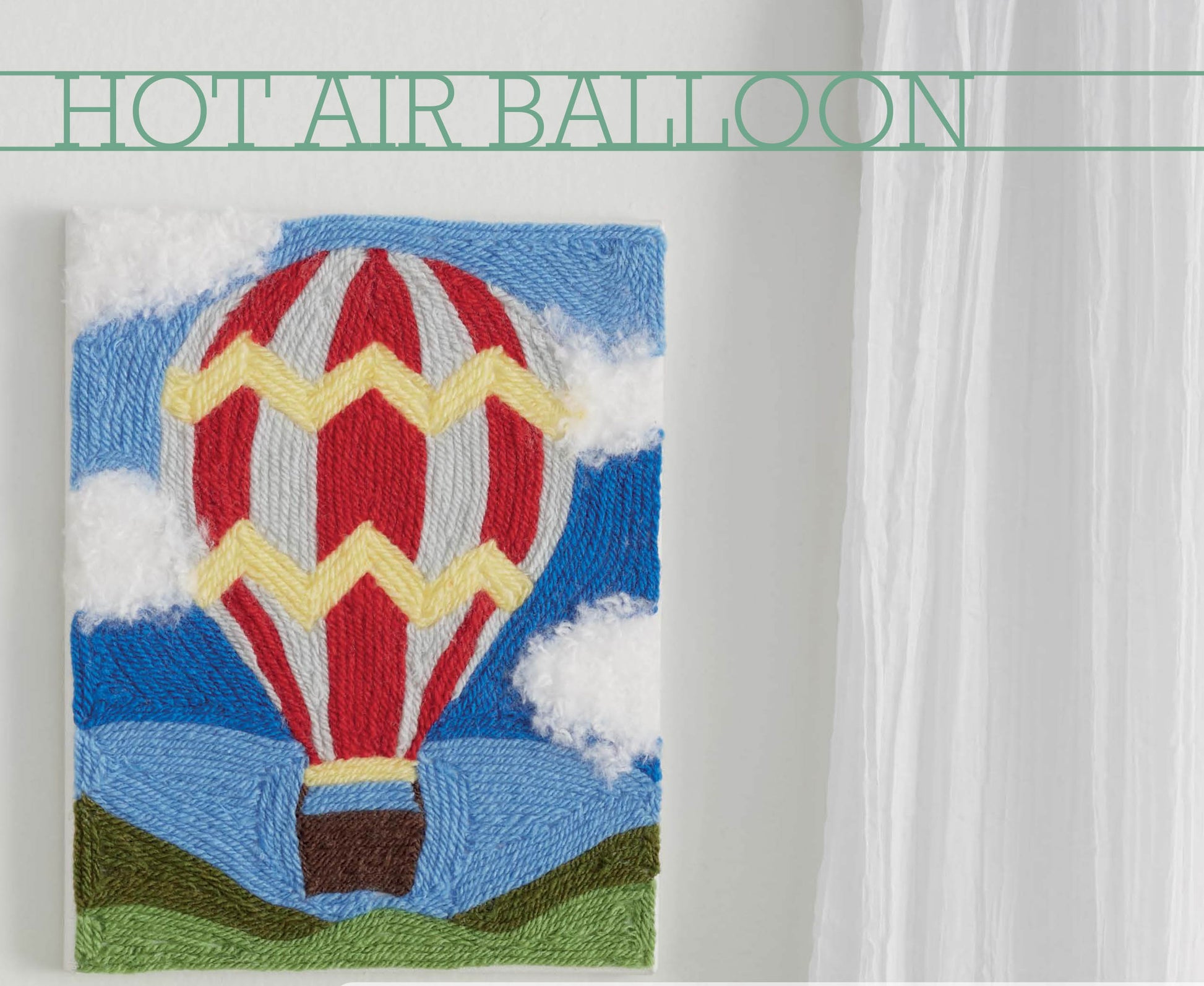 The Yarn Painting Digital Download by Leisure Arts features a hot air balloon with red, yellow, and gray stripes floating above green hills—ideal for beginners or creative canvas projects. Displayed on a white wall near sheer curtains. Text reads HOT AIR BALLOON.