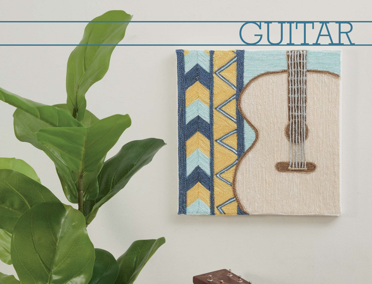 The Yarn Painting Digital Download by Leisure Arts displays a small guitar art piece with colorful geometric canvas patterns on the left. “GUITAR” appears in blue at the top right. It’s shown hanging on a white wall near a leafy green plant.
