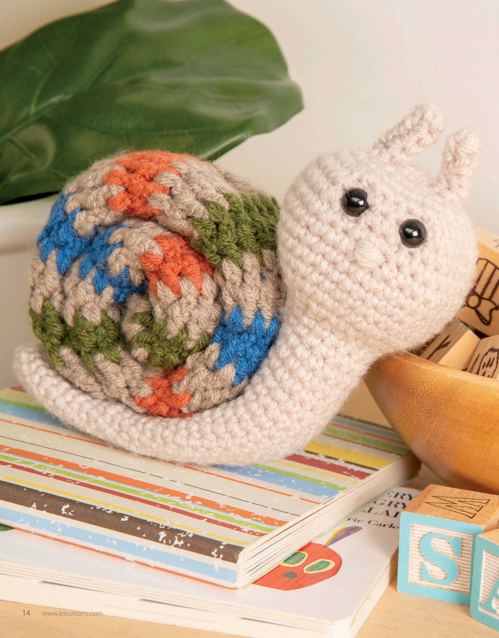 The Mini Huggables Crochet snail plush by Leisure Arts, inspired by amigurumi woodland animals, sits on children's books beside wooden alphabet blocks, with a plant in the background.