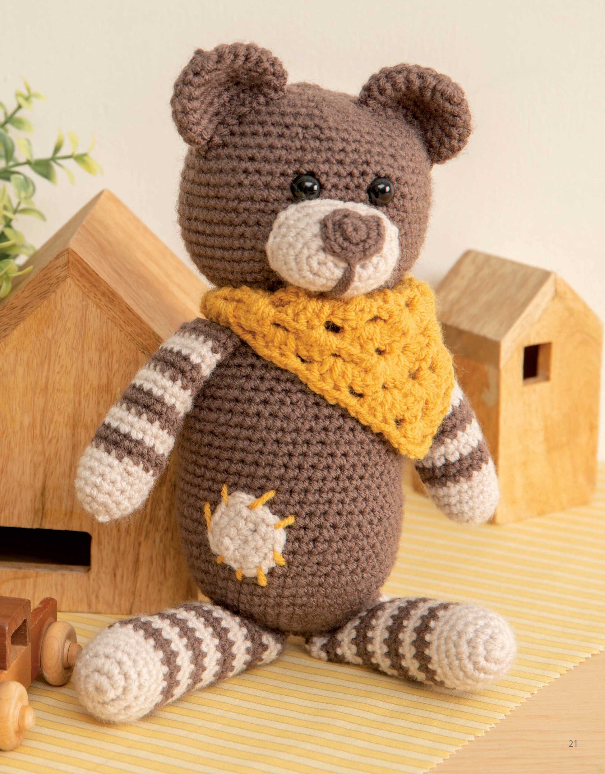 The Mini Huggables Crochet bear by Leisure Arts, featuring a yellow scarf and sun patch, sits on a striped mat surrounded by wooden toy houses and a plant.
