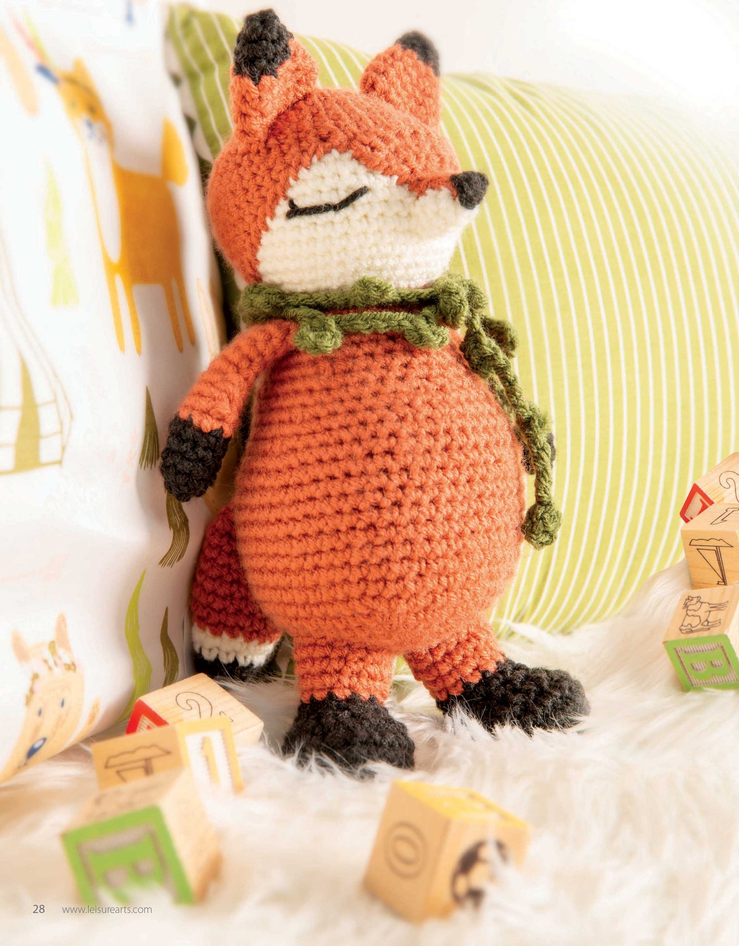 The Mini Huggables Crochet fox plush by Leisure Arts, crafted using beginner patterns, stands among wooden alphabet blocks on a fluffy white surface. Its closed eyes, green scarf, and black-tipped ears capture classic amigurumi woodland charm.
