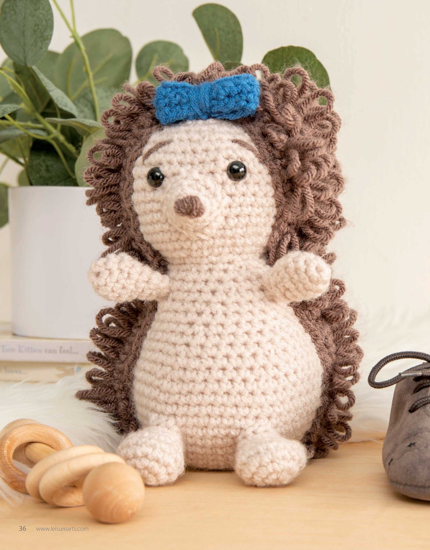 The Mini Huggables Crochet hedgehog by Leisure Arts, featuring curly brown yarn spikes, a beige body, black eyes, and a blue bow, sits on a table beside a plant, wooden rattle, and shoe.