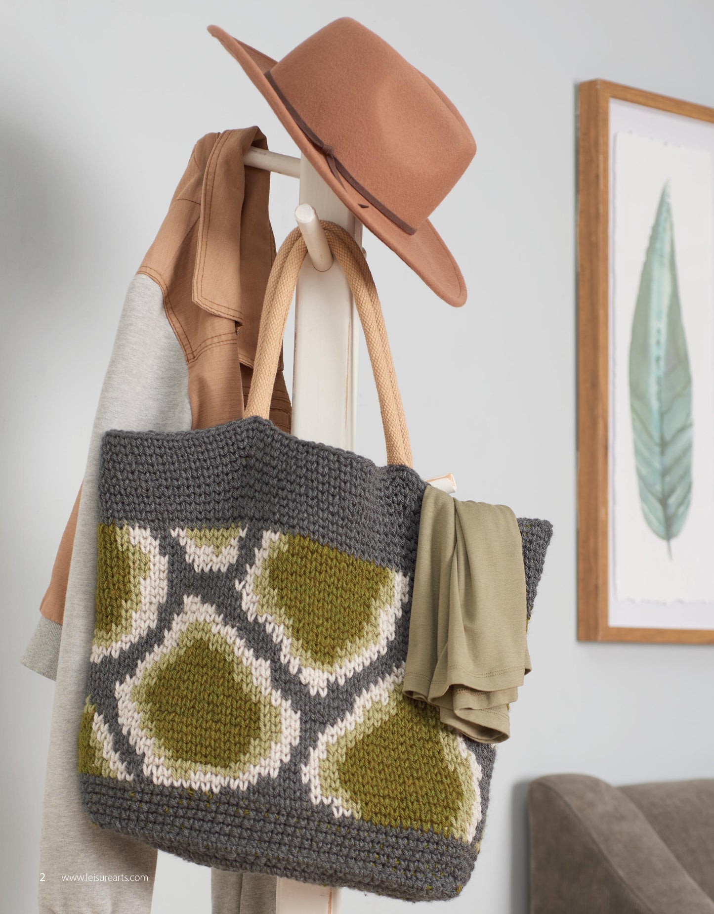 The knit tote bag features a geometric pattern inspired by "Animal Prints—10 Designs to Add a Little Wildlife to Your Wardrobe and Home Decor" by Leisure Arts. It hangs on a white coat rack with stylish accessories, with feather artwork in the background.