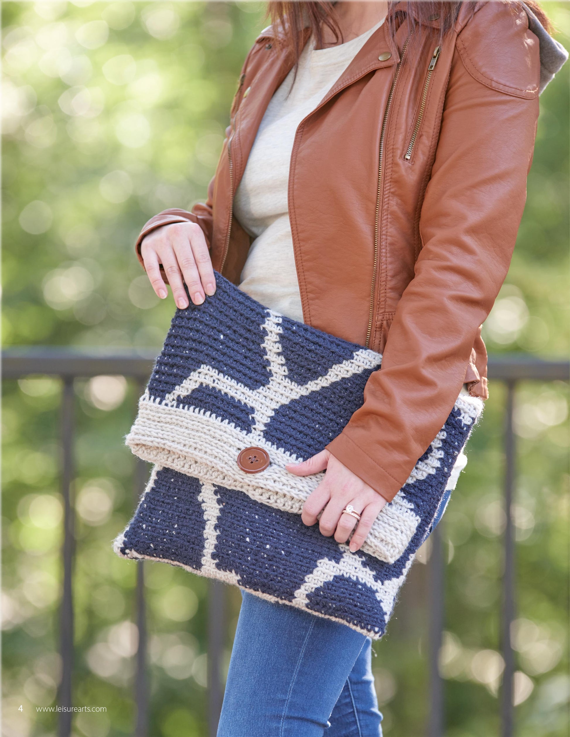 A person in a brown leather jacket and blue jeans holds Leisure Arts' "Animal Prints—10 Designs to Add a Little Wildlife to Your Wardrobe and Home Decor" outdoors, with a crocheted clutch featured against a blurred green background.