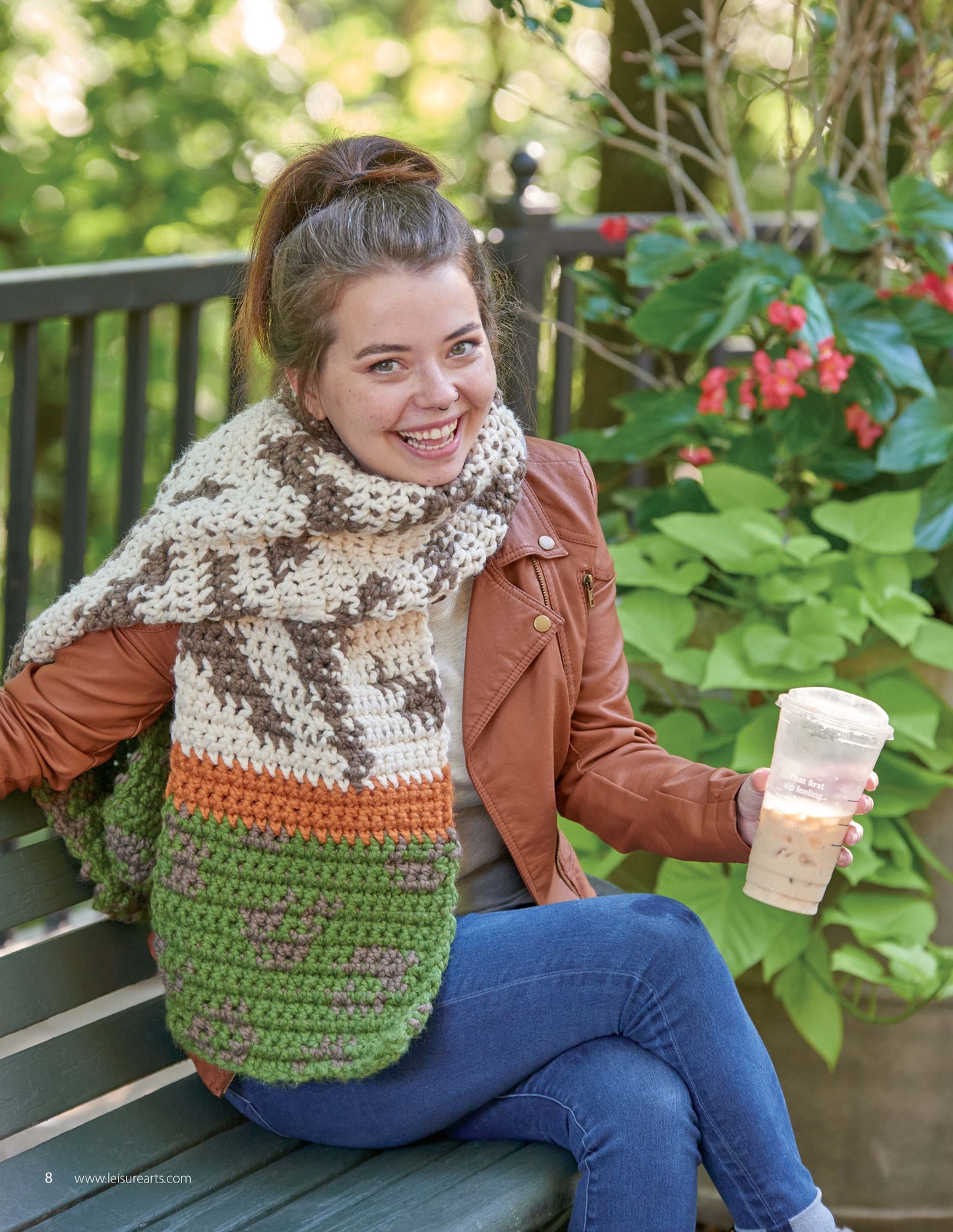 A smiling woman with a ponytail sits on a park bench, wearing a brown jacket and a chunky crochet scarf. Her look is perfect inspiration for creating animal-themed pieces from "Animal Prints—10 Designs" by Leisure Arts.