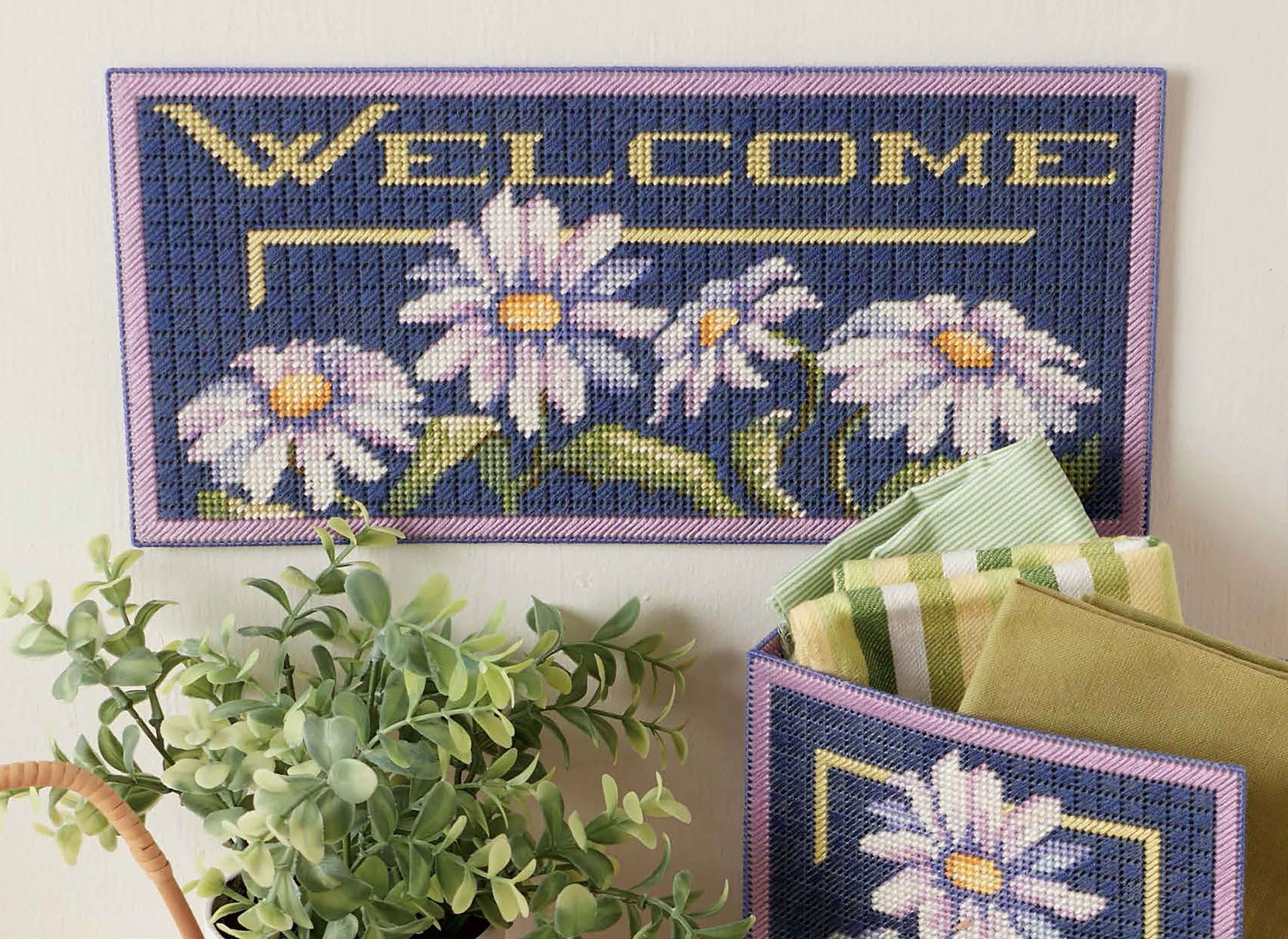The Lazy Daisies of Summer wall hanging by Leisure Arts features "WELCOME" and white daisies on blue, with a matching pocket for cloths—ideal beginner home decor for embroidery floss craft lovers, displayed beside a potted plant.