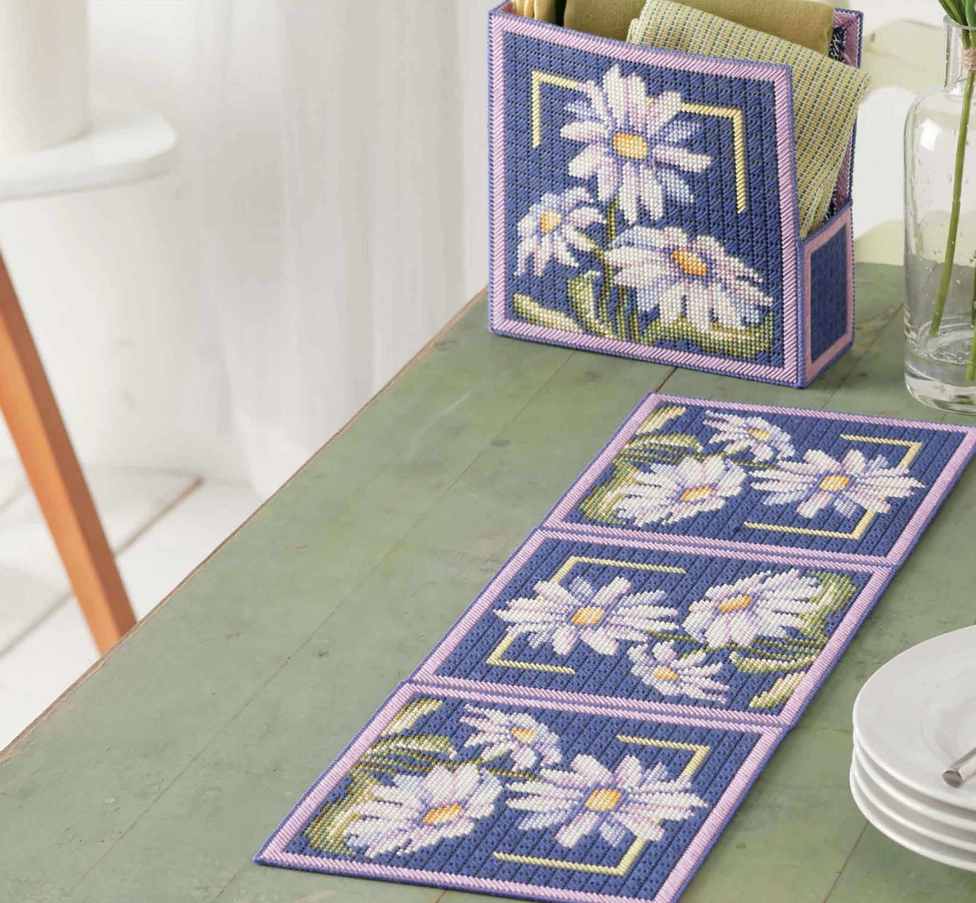 The Leisure Arts "Lazy Daisies of Summer" set features a plastic canvas napkin holder and matching table runner for beginners, both decorated with white daisies, yellow centers, green stems, and purple-trimmed blue backgrounds.