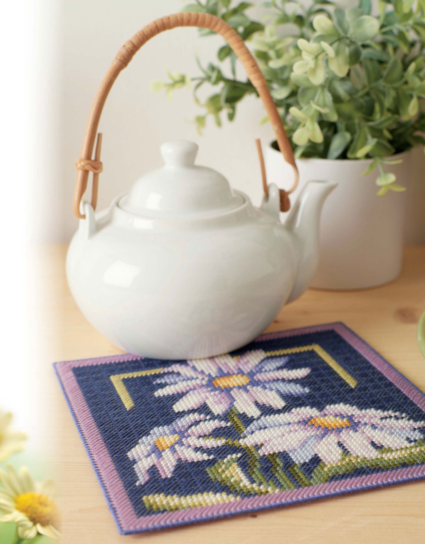 The "Lazy Daisies of Summer" by Leisure Arts features a white teapot with a curved handle on a wooden table, set atop a floral-patterned mat, with a green potted plant and yellow flowers softly blurred in the background.