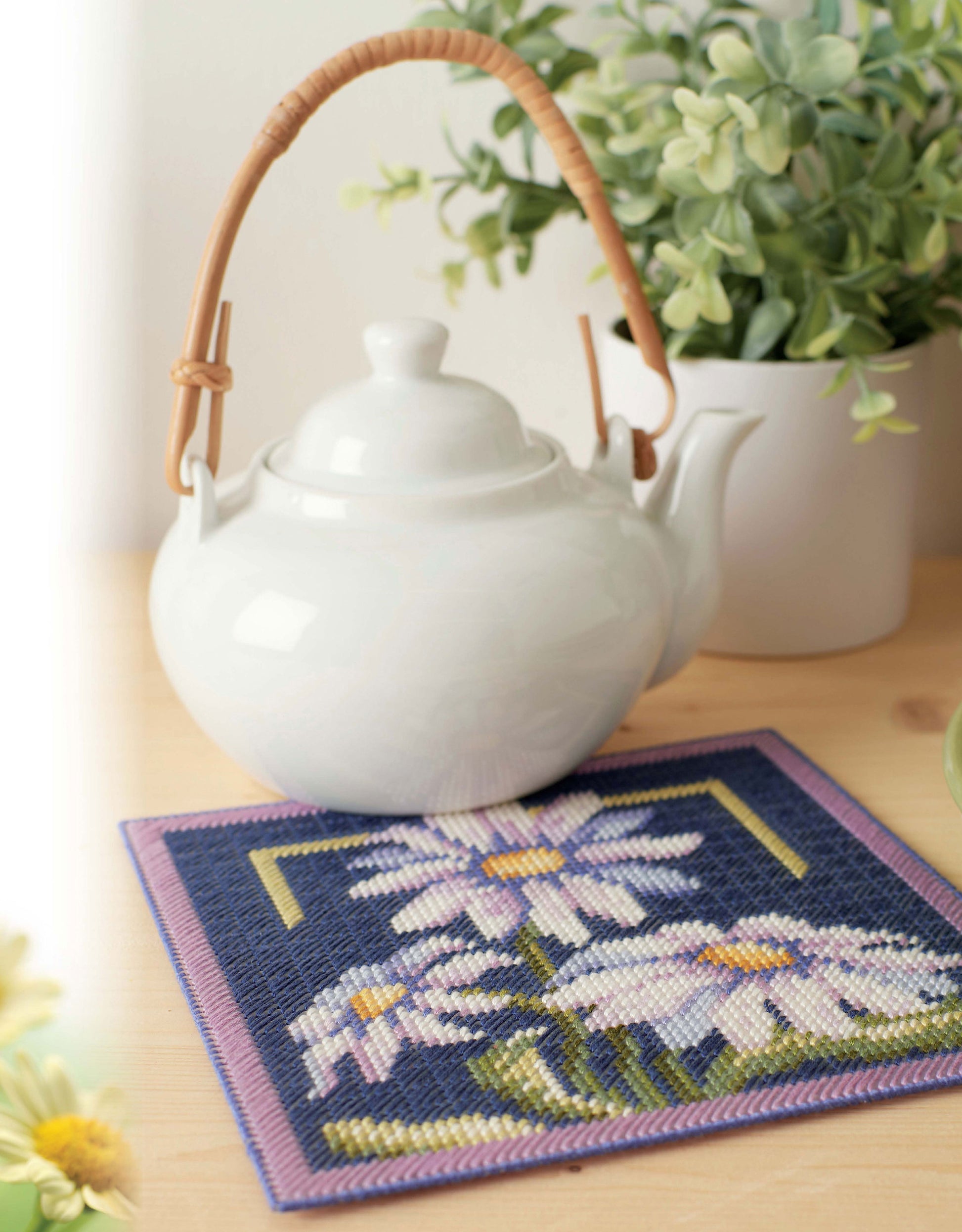 The "Lazy Daisies of Summer" by Leisure Arts features a white teapot with a curved handle on a wooden table, set atop a floral-patterned mat, with a green potted plant and yellow flowers softly blurred in the background.