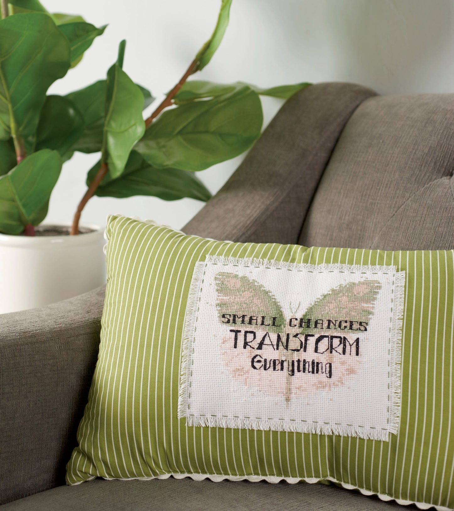 A pillow with "SMALL CHANGES TRANSFORM" from Leisure Arts' Positive Quotes in Cross Stitch Digital Download rests on a gray armchair next to a potted plant with large green leaves.