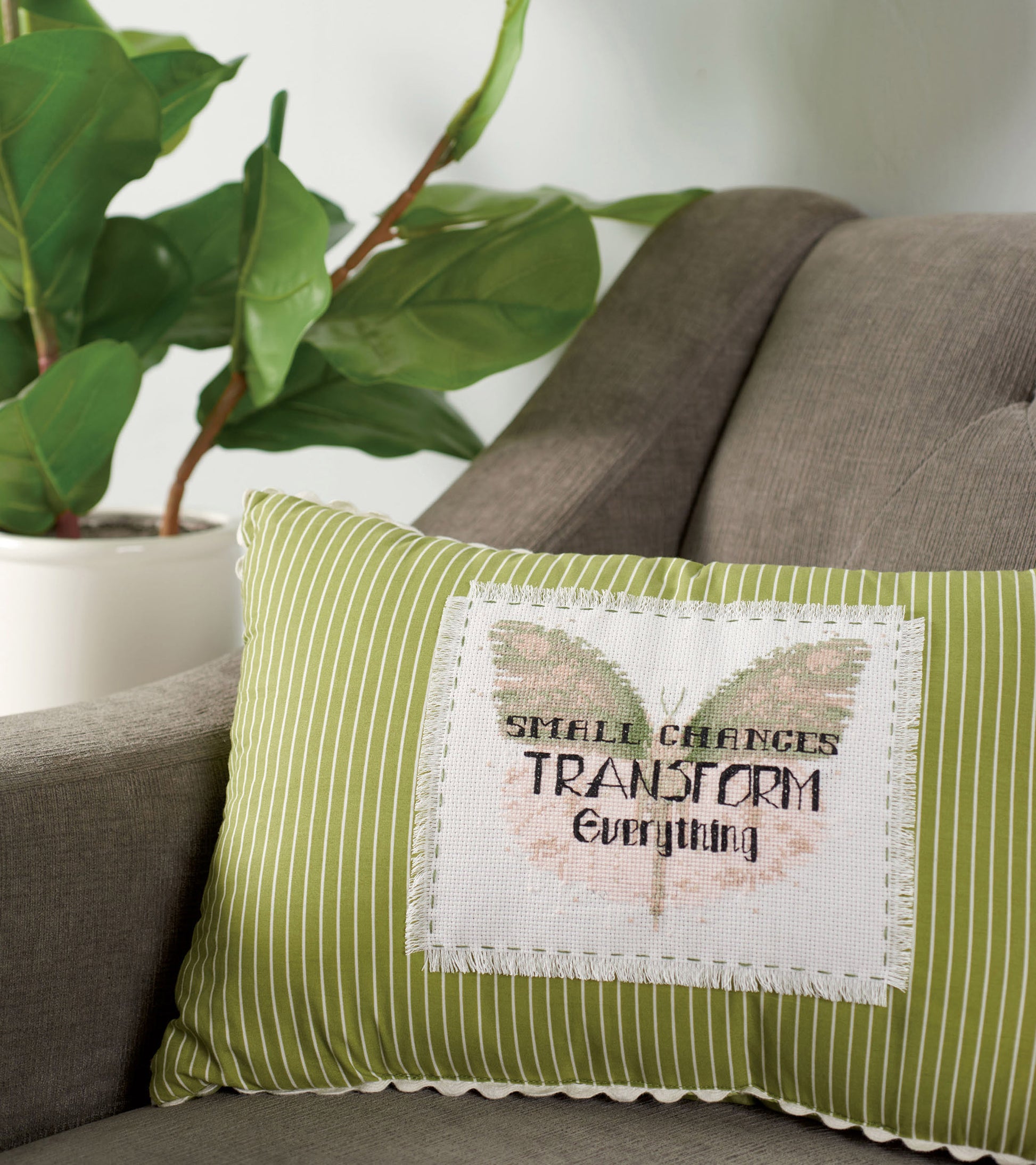 A pillow with "SMALL CHANGES TRANSFORM" from Leisure Arts' Positive Quotes in Cross Stitch Digital Download rests on a gray armchair next to a potted plant with large green leaves.