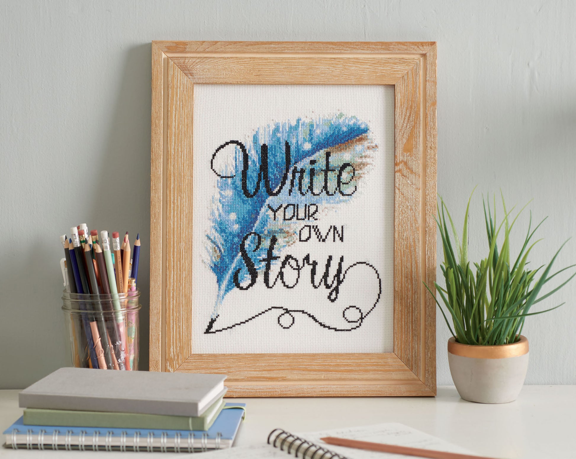 The Positive Quotes in Cross Stitch Digital Download by Leisure Arts features “Write your own story” in black script with a blue feather. Ideal for beginners, it’s shown with colored pencils, notebooks, and a potted plant.
