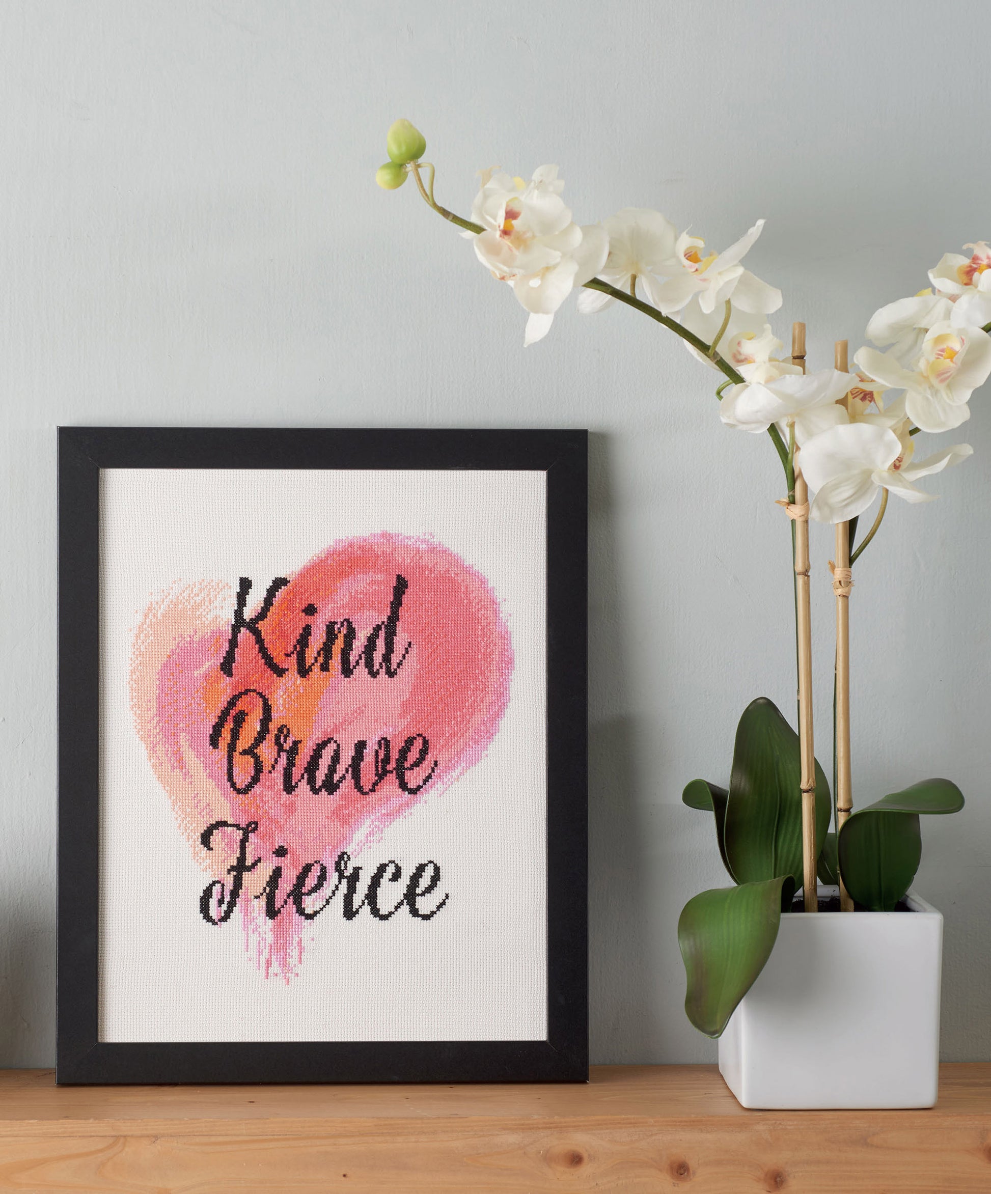The Positive Quotes in Cross Stitch Digital Download by Leisure Arts features the words Kind Brave Fierce over a pink watercolor heart, displayed on a wooden surface beside a white orchid in bloom.