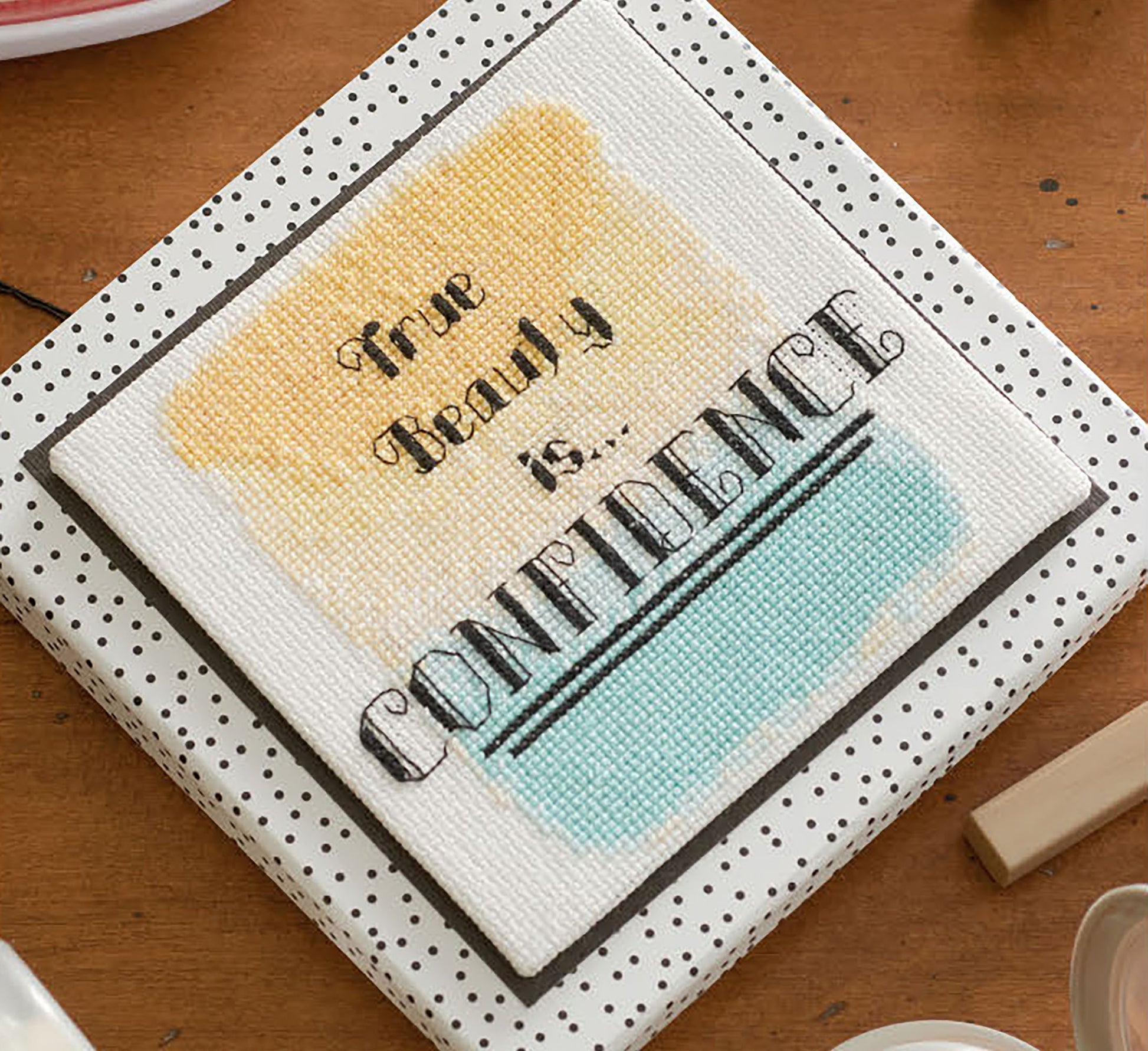 Leisure Arts’ Positive Quotes in Cross Stitch Digital Download features “True beauty is... CONFIDENCE” on a white canvas with yellow and blue watercolor backgrounds, ideal for beginners and shown on a polka-dotted surface.