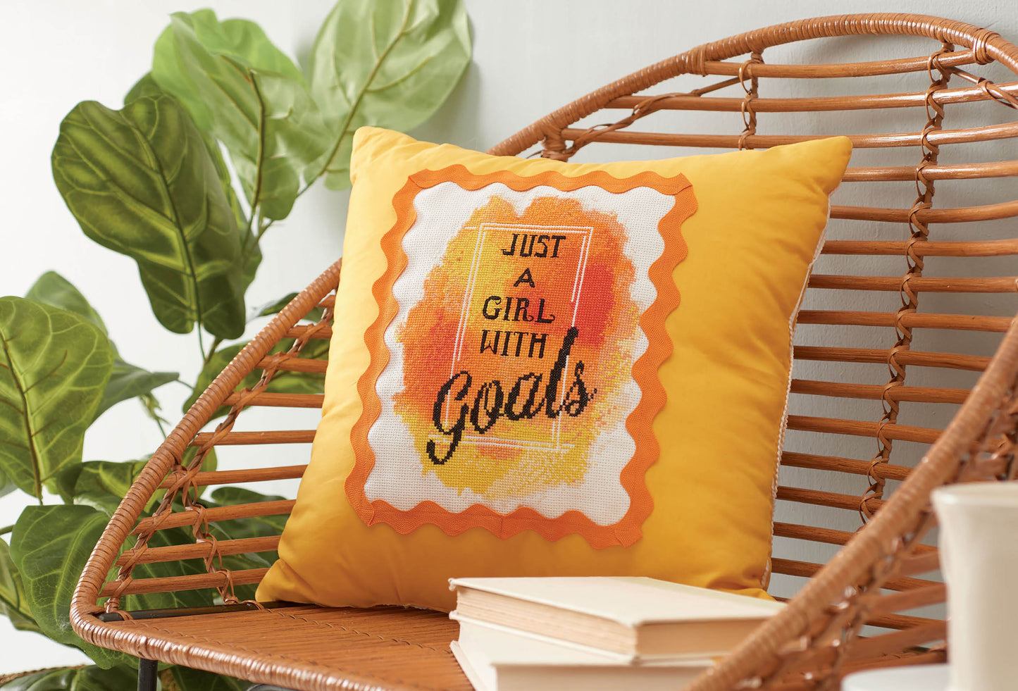 A yellow throw pillow on a wicker chair displays "Just a girl with goals," made with the Leisure Arts Positive Quotes in Cross Stitch Digital Download. This cozy nook also features a green plant and an inspiring book stack.