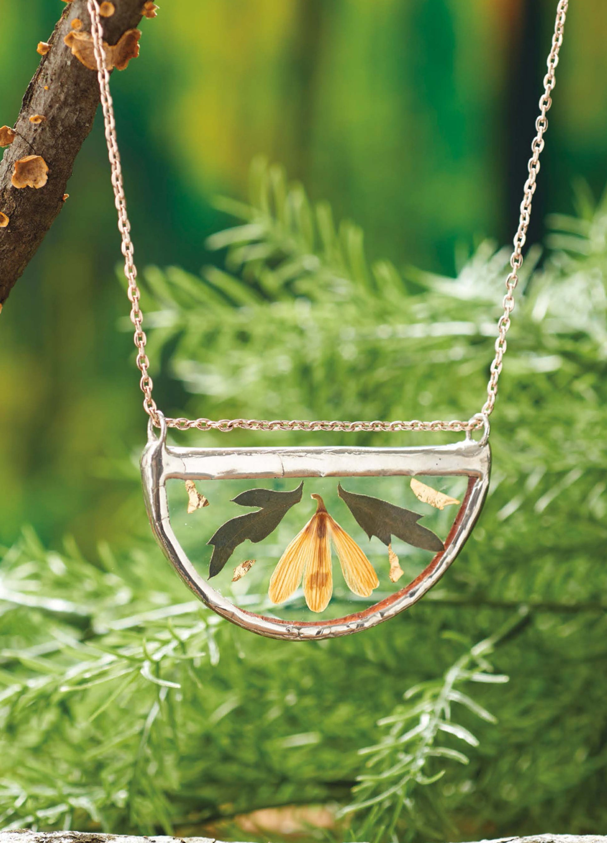 A close-up of the "Stained Glass for Beginners Digital Download" by Leisure Arts shows a semicircular glass art pendant with dried leaves and a maple seed, set against blurred greenery with tree branches and pine needles.