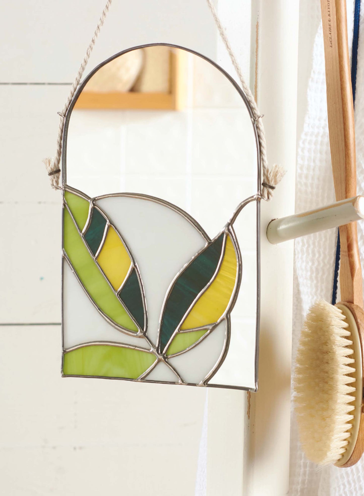 The Stained Glass for Beginners Digital Download by Leisure Arts features a small leaf design in green, yellow, and white—perfect for starter projects—shown hanging by a rope on a door handle beside a wooden bath brush and white towel.