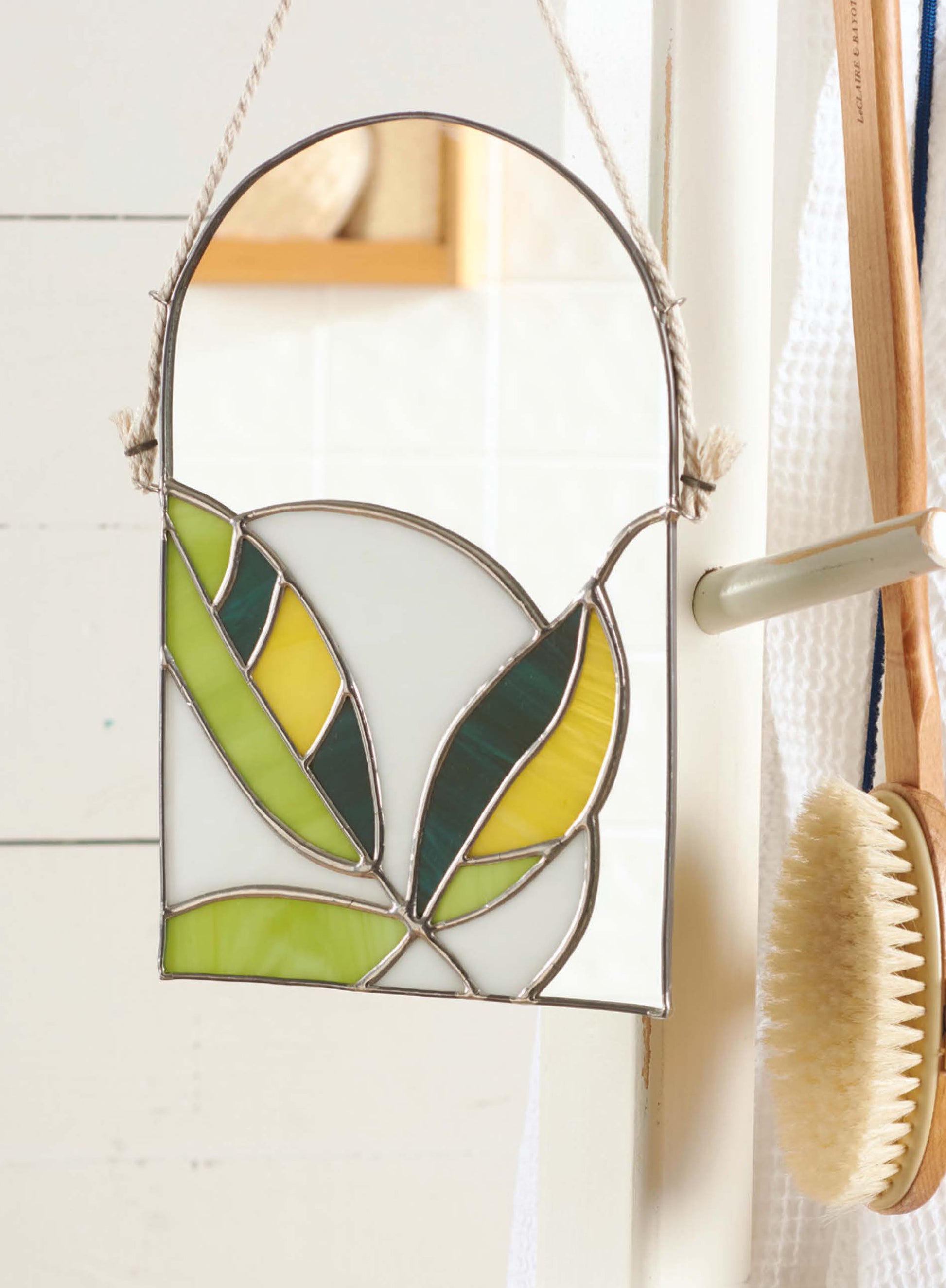 The Stained Glass for Beginners Digital Download by Leisure Arts features a small leaf design in green, yellow, and white—perfect for starter projects—shown hanging by a rope on a door handle beside a wooden bath brush and white towel.