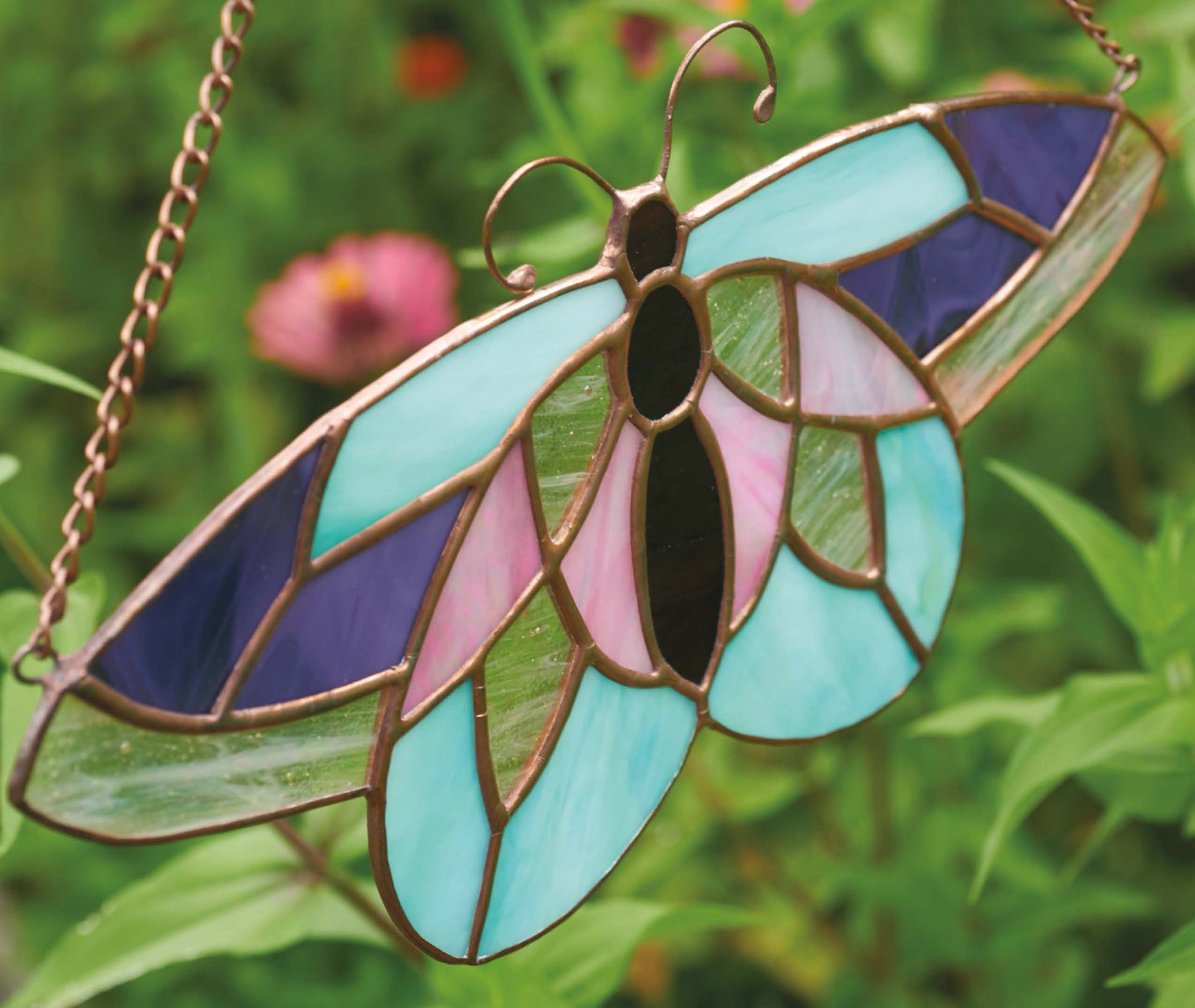 The Stained Glass for Beginners Digital Download by Leisure Arts features a butterfly ornament with blue, purple, pink, and green wings. It hangs outdoors on a metal chain with blurred greenery and a pink flower in the background.