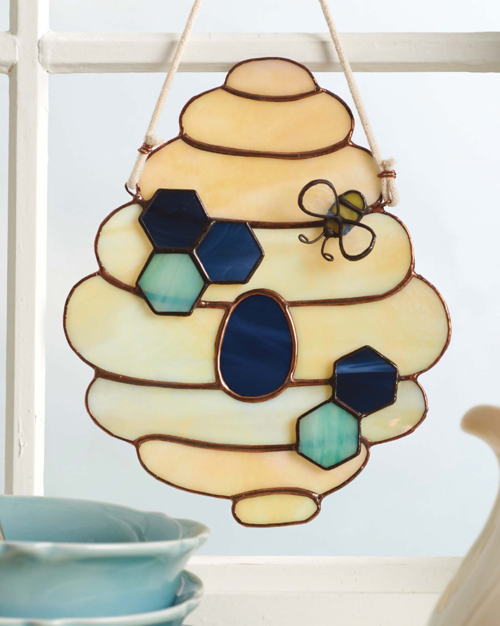 The "Stained Glass for Beginners Digital Download" by Leisure Arts features a beehive-shaped art piece with blue and teal hexagons, an oval, and a wire bee—ideal for starter projects. Blue dishes appear below the window display.