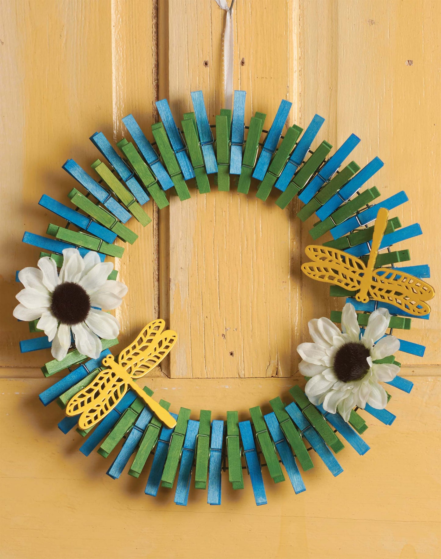 The Leisure Arts Clothespin Wreaths DIY kit creates a blue and green wreath with white faux flowers and yellow dragonflies, perfect for home decor projects. Shown displayed on a light yellow wooden door.