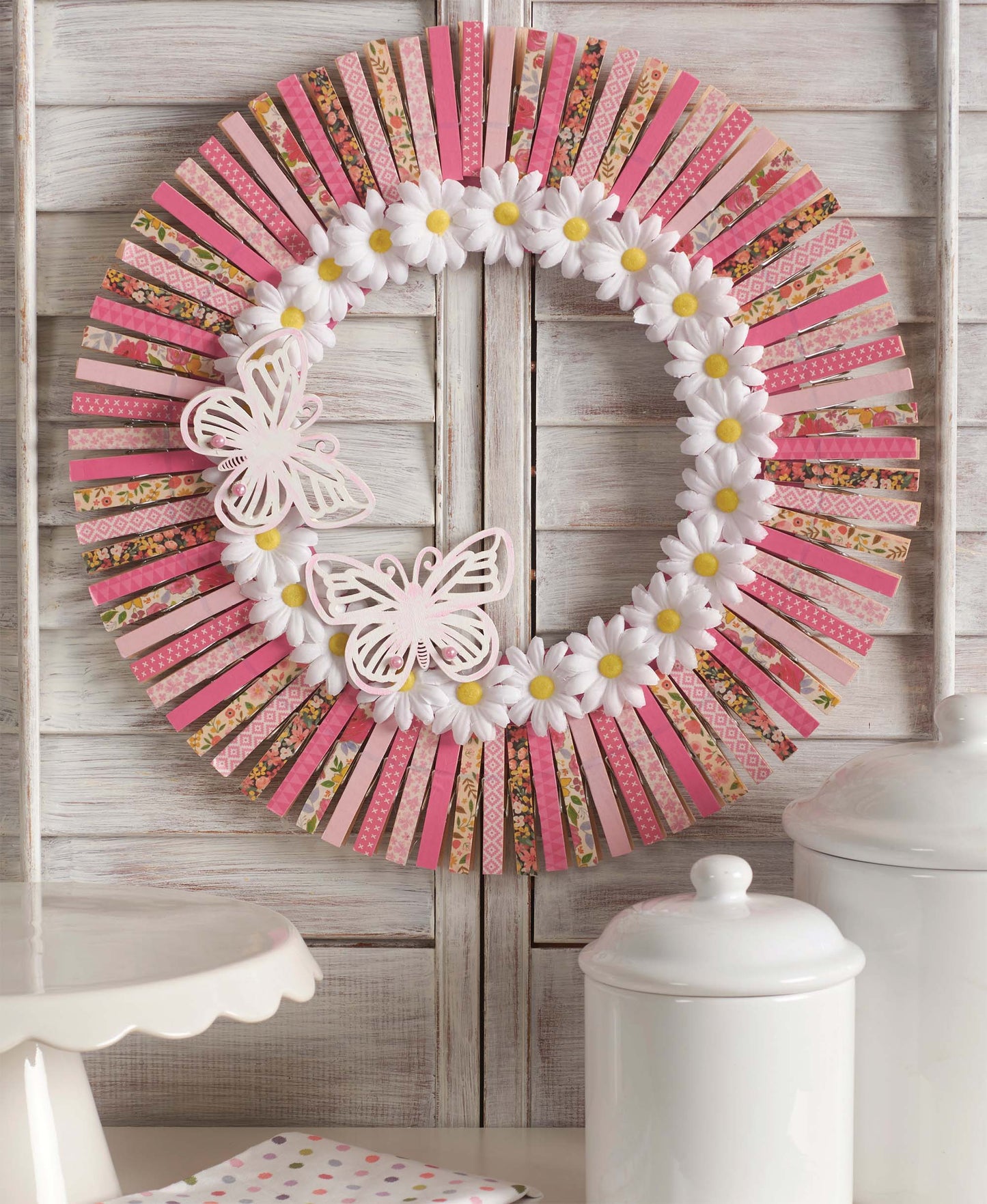 The Leisure Arts Clothespin Wreaths kit features pink, red, and patterned clothespins with white daisies and butterfly ornaments—a charming DIY decor project perfect for adding a seasonal touch to your home.