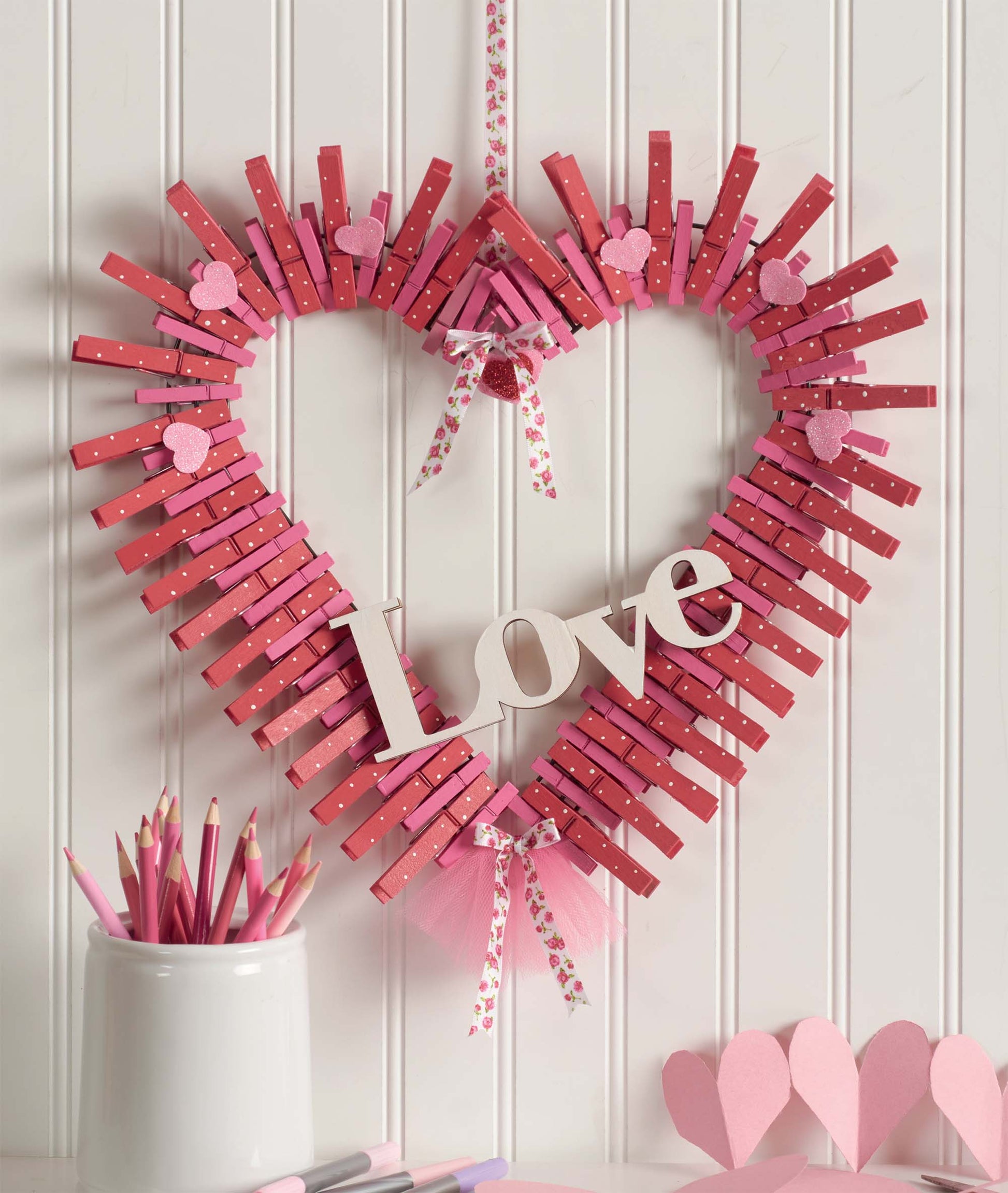 The Clothespin Wreaths by Leisure Arts features a heart-shaped design made with pink clothespins, bows, and hearts. "Love" is in the center, with pink pencils and heart paper in a white cup—ideal for DIY enthusiasts.