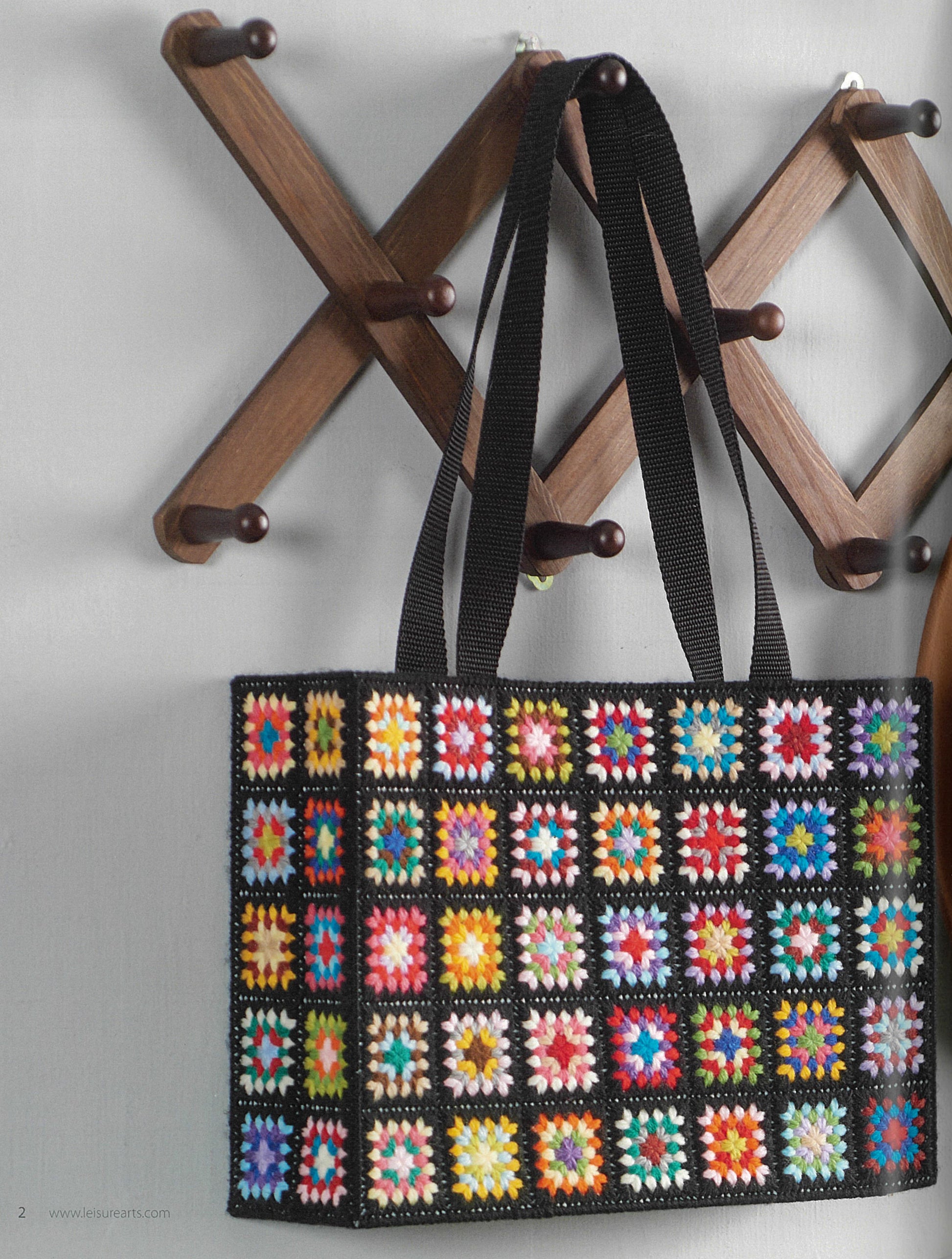 The Granny Square Tote & Wallet Digital Download by Leisure Arts features a colorful retro crochet tote with black handles, shown hanging on a wooden peg rack against a light gray wall.