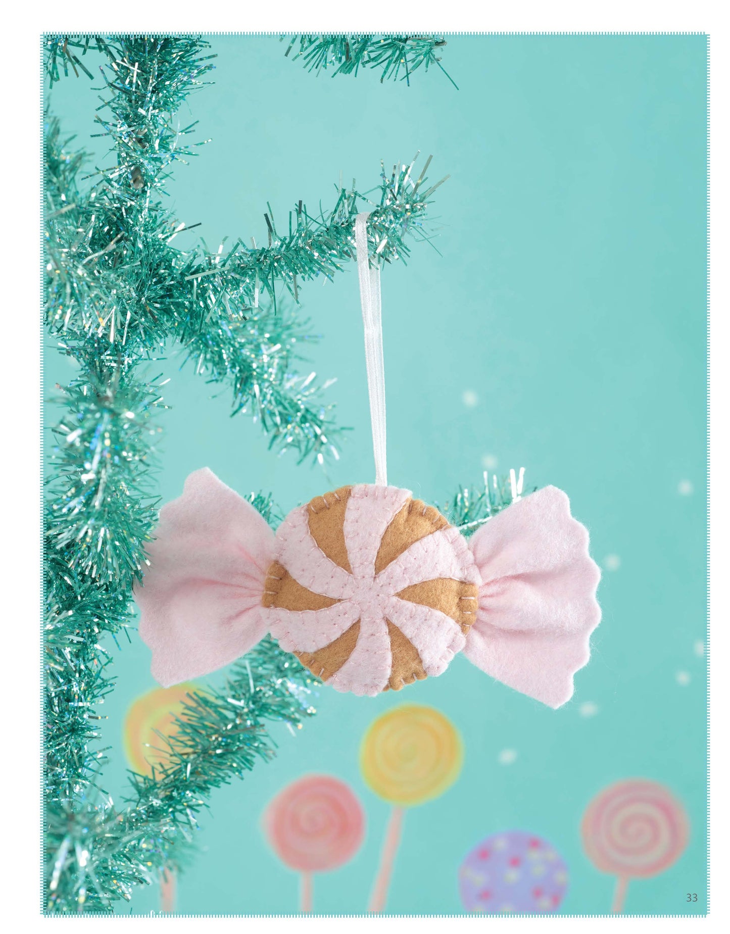 The Gingerbread House ornament by Leisure Arts, perfect for wool felt projects, hangs from a teal tinsel Christmas tree. It features handmade felt details in pink and gold with pastel decorations resembling a festive candy scene.