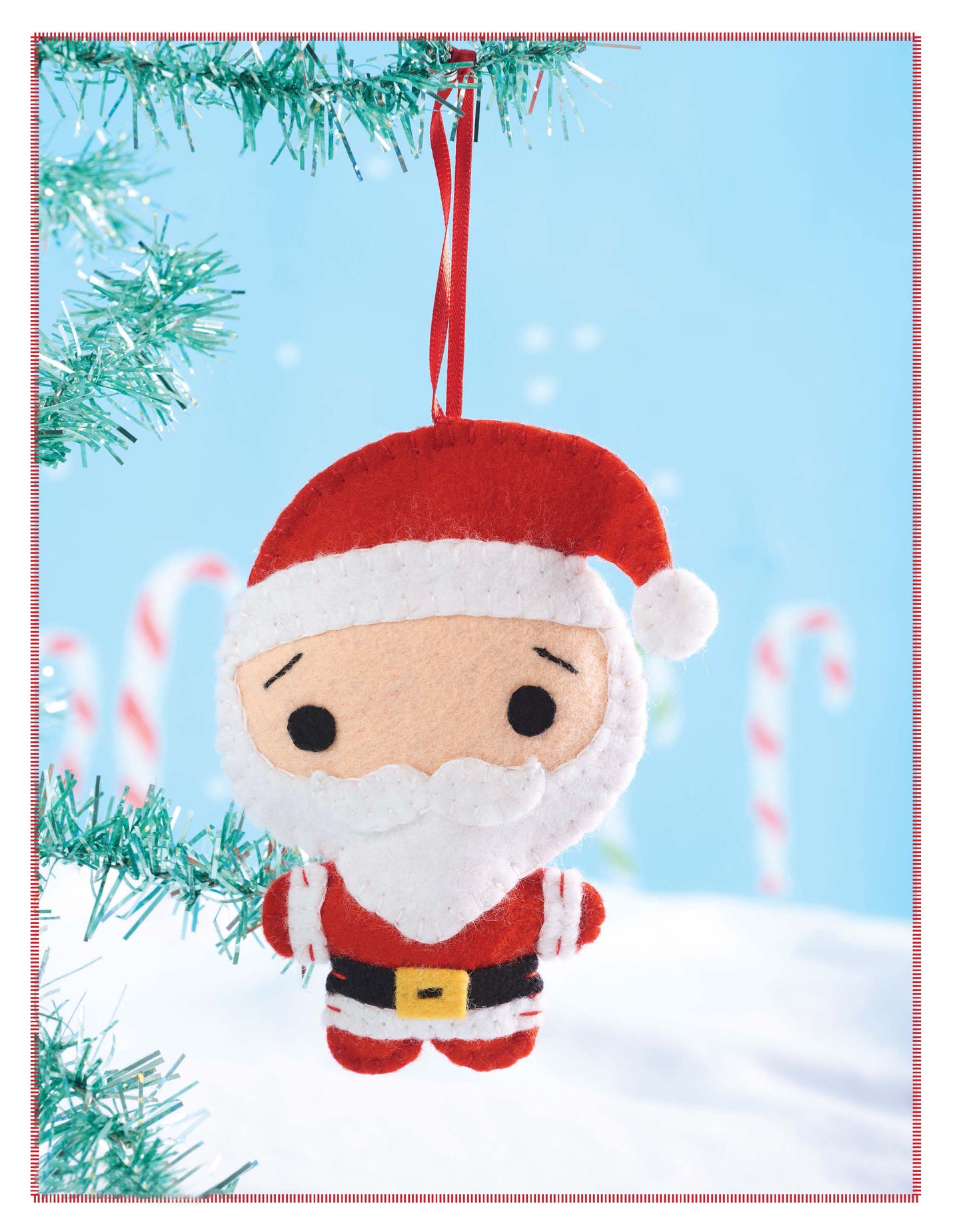 Santa's Workshop by Leisure Arts features a cute felt Santa Claus with a white beard, hanging from a red ribbon on a green tree branch. Snow and candy canes decorate the scene under a bright blue sky.