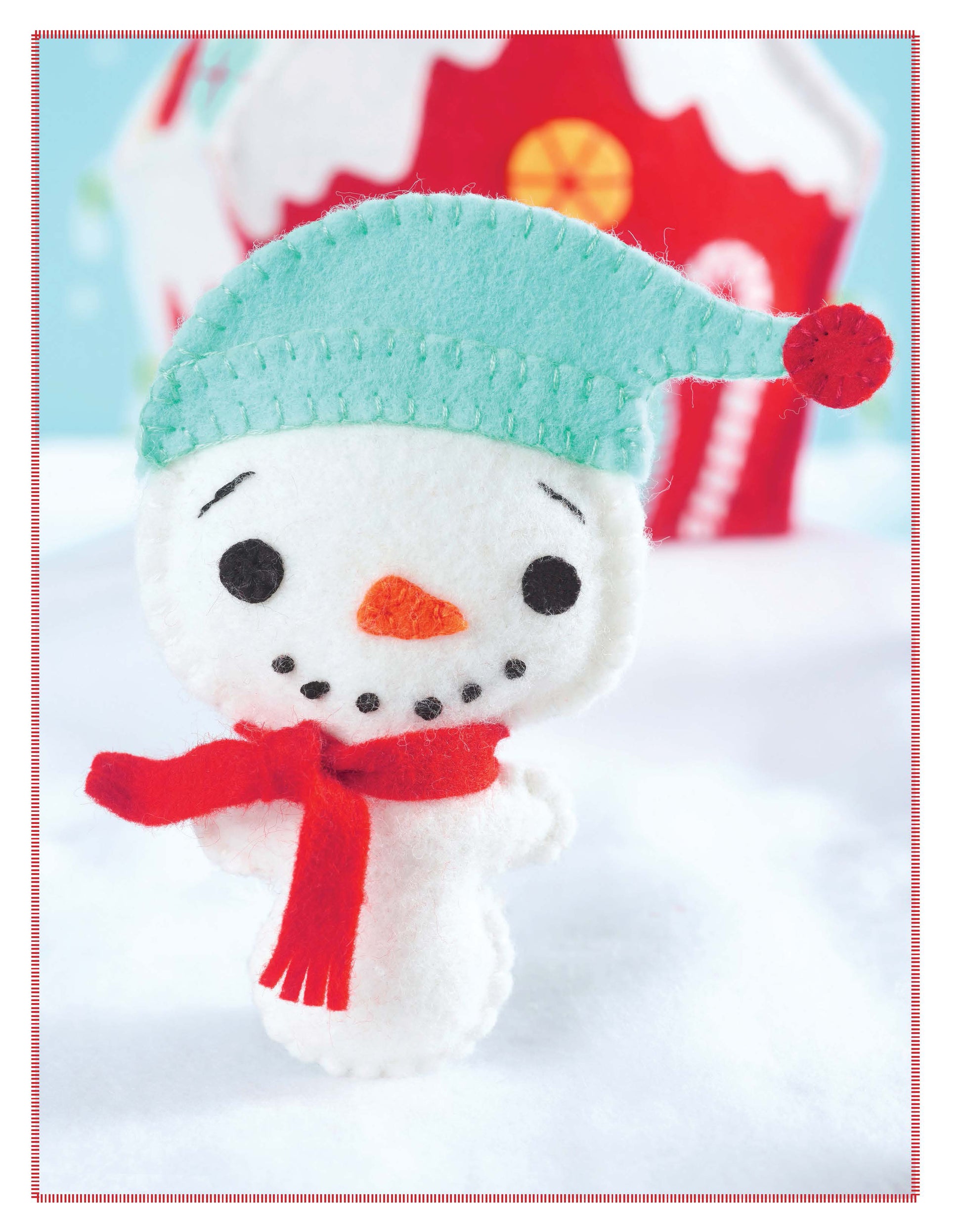 Santa's Workshop by Leisure Arts is a handmade felt snowman wearing a turquoise hat and red scarf, with black stitched eyes and mouth. He stands in snowy scenery with a blurred red and white house behind him.
