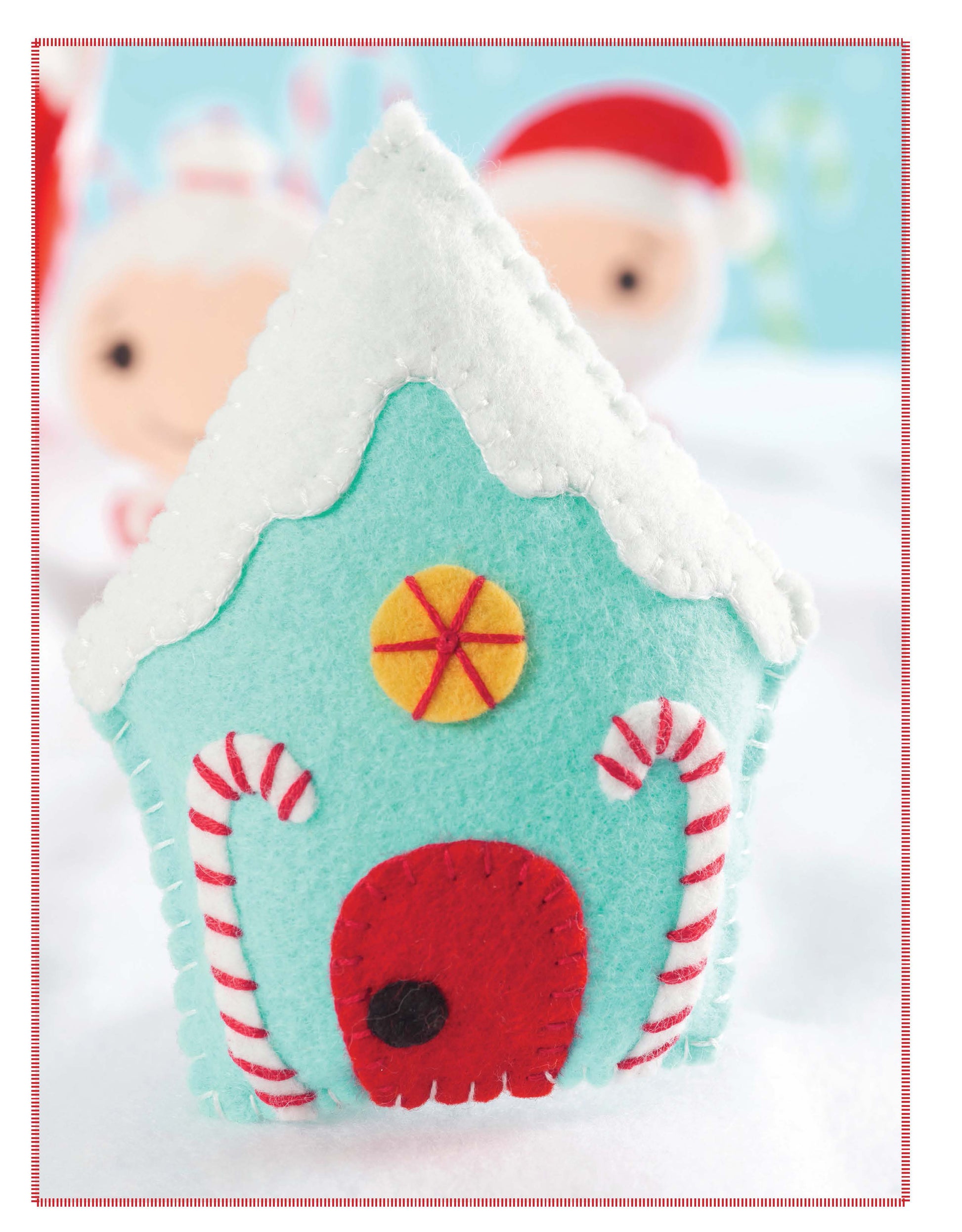 Santa’s Workshop by Leisure Arts is a small, light blue felt house with a white snow-like roof, red felt door, candy cane accents, and a yellow circle above the door—perfect for Christmas felt crafts; blurred plush figures in the background.
